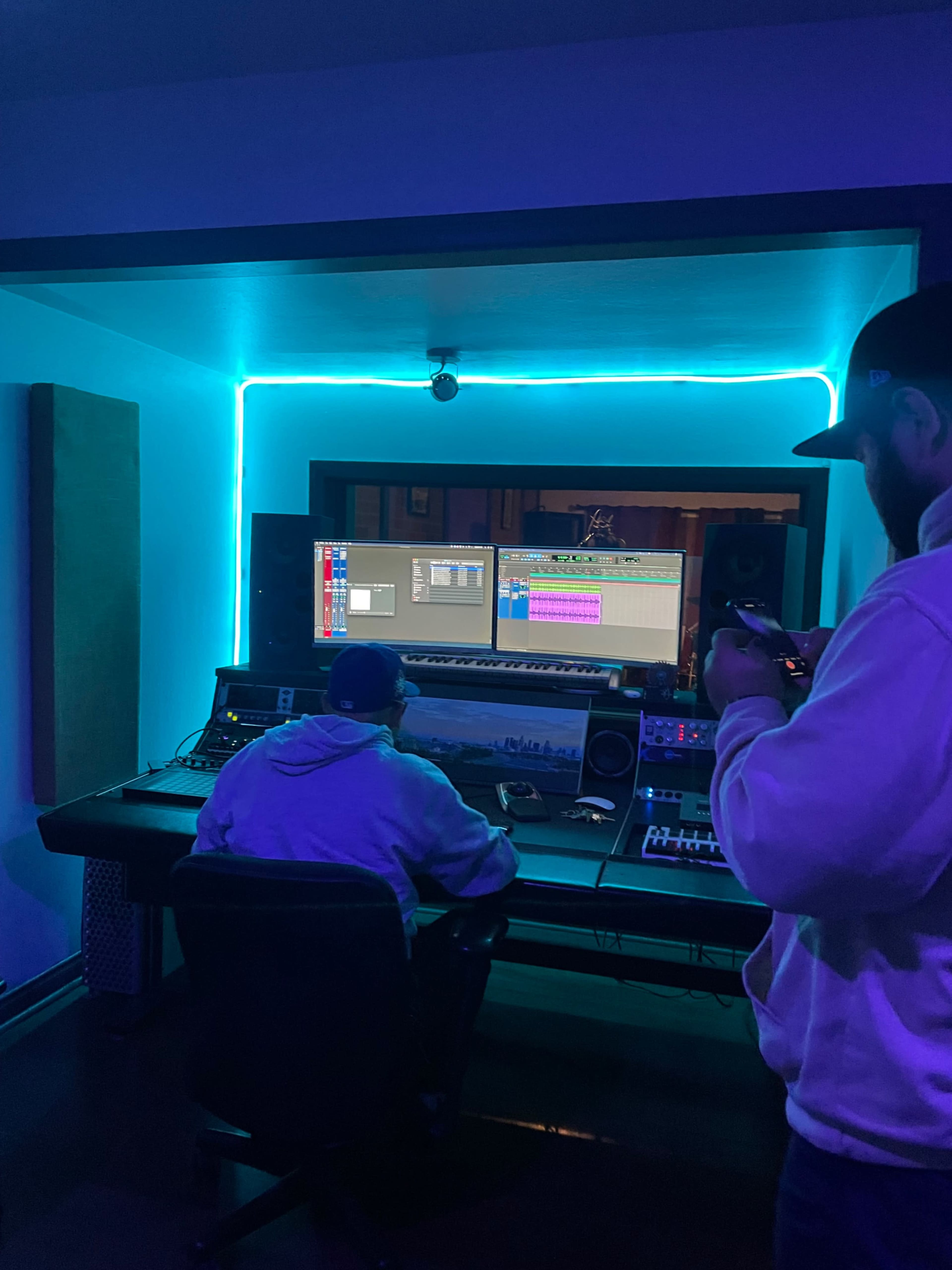 A person sits at a recording studio desk while another person stands nearby, holding a phone, in a room illuminated by blue neon lighting.