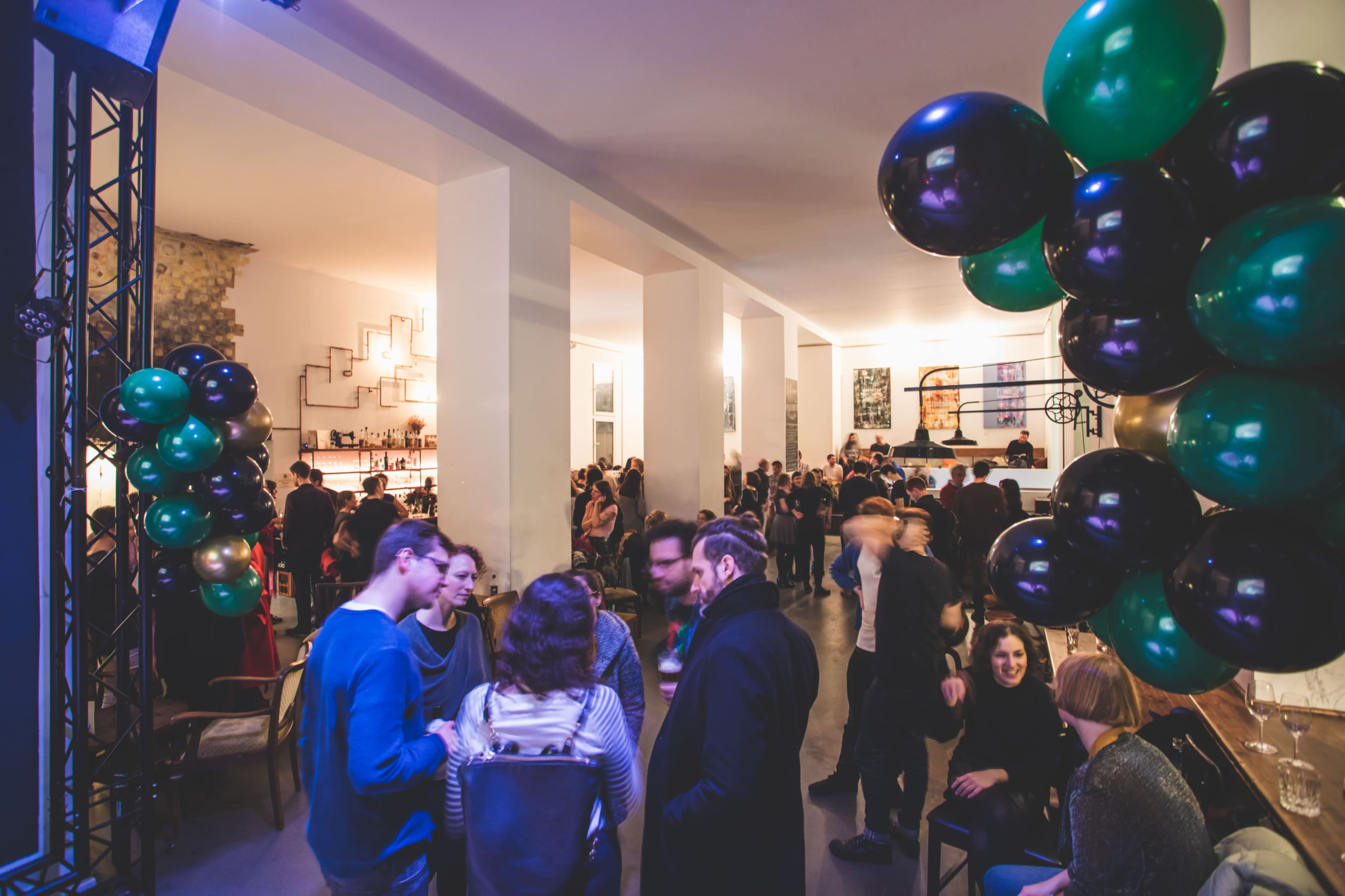 A crowd of people socializes at a lively indoor party, surrounded by decorations including large green and black balloons.