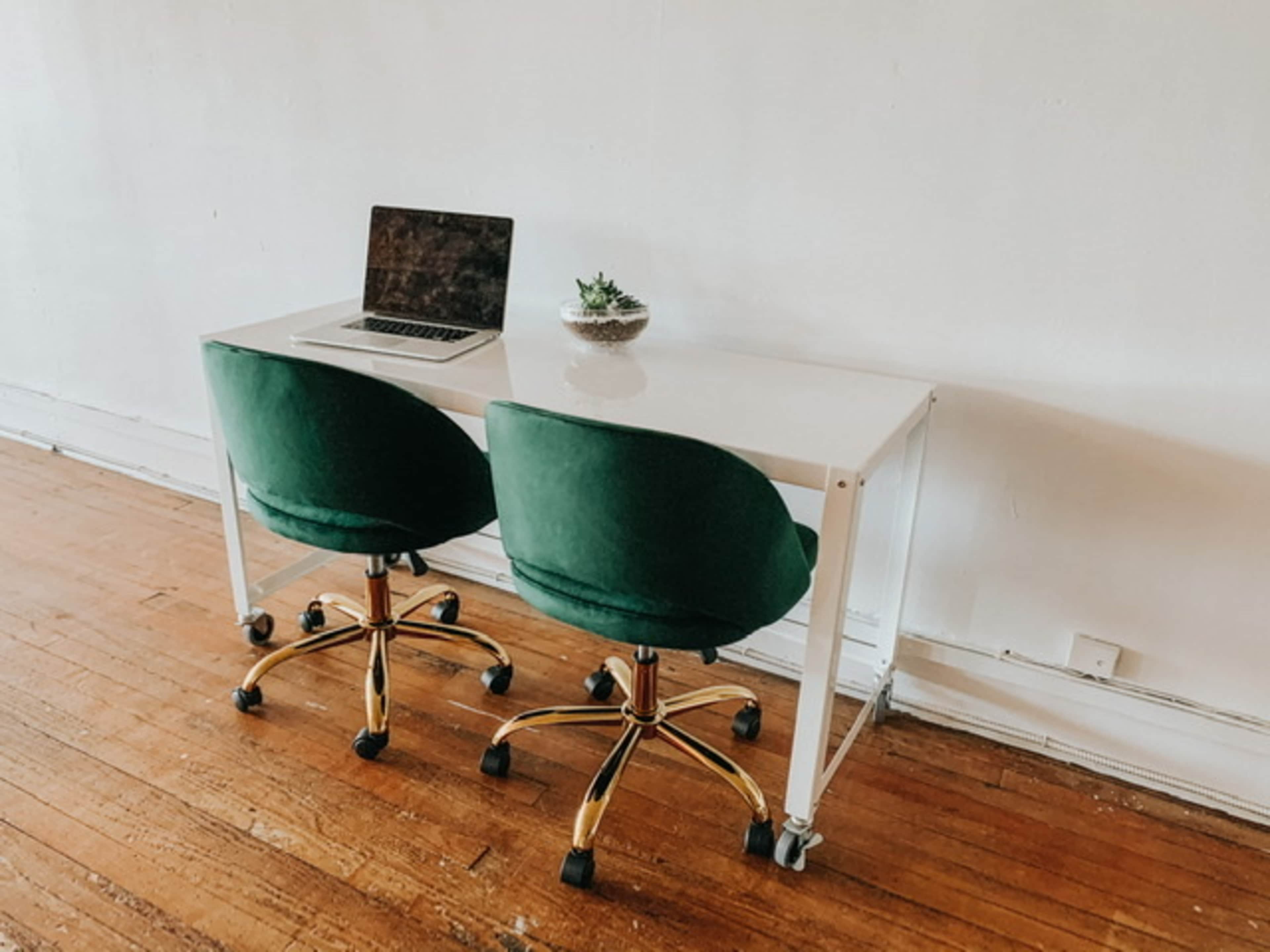 A modern desk with a laptop and two green chairs on wheels is positioned against a plain white wall.