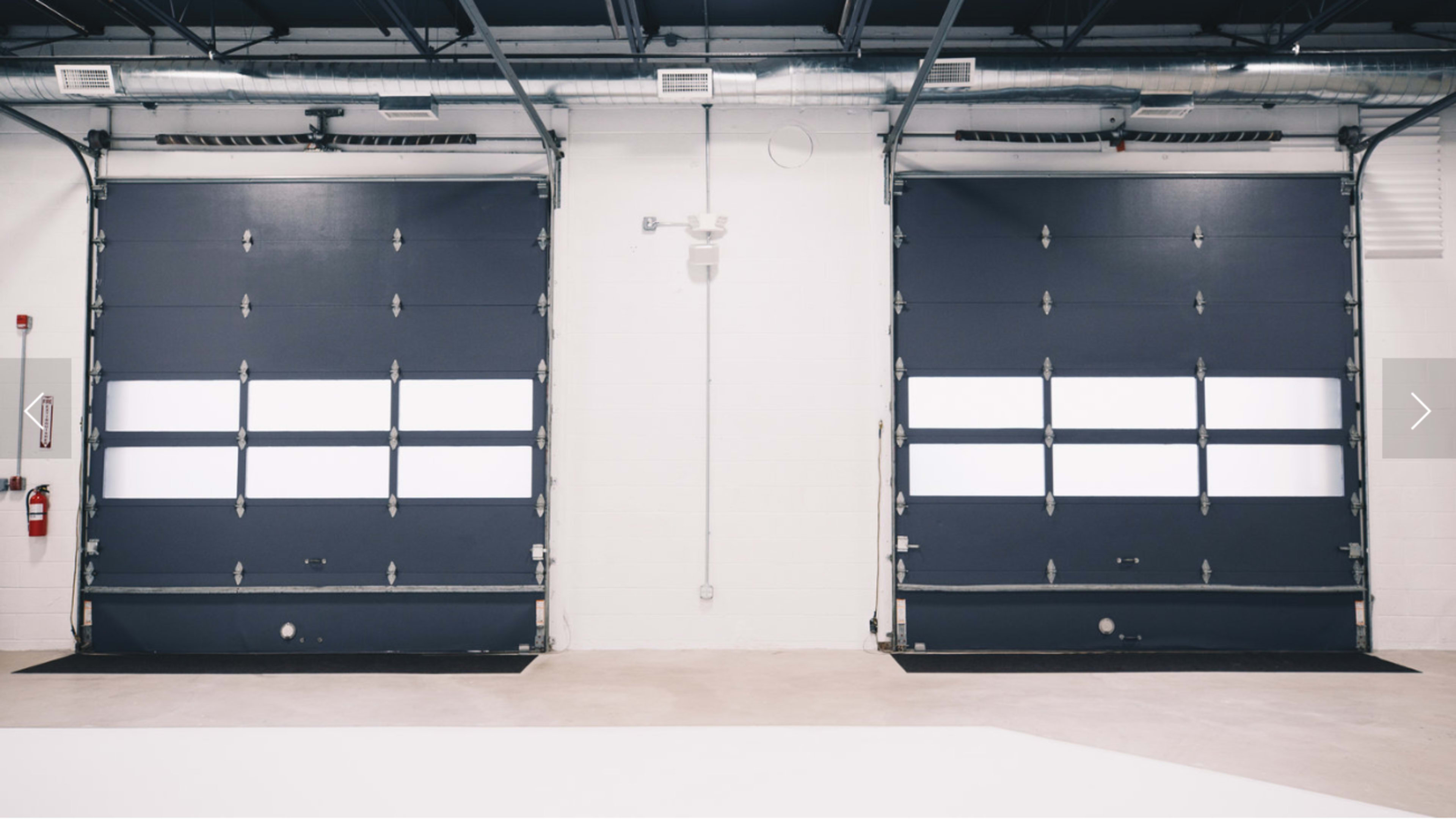 The image shows a spacious industrial area with two large dark blue garage doors on the front wall, each featuring horizontal windows, and a clean, uncluttered floor.
