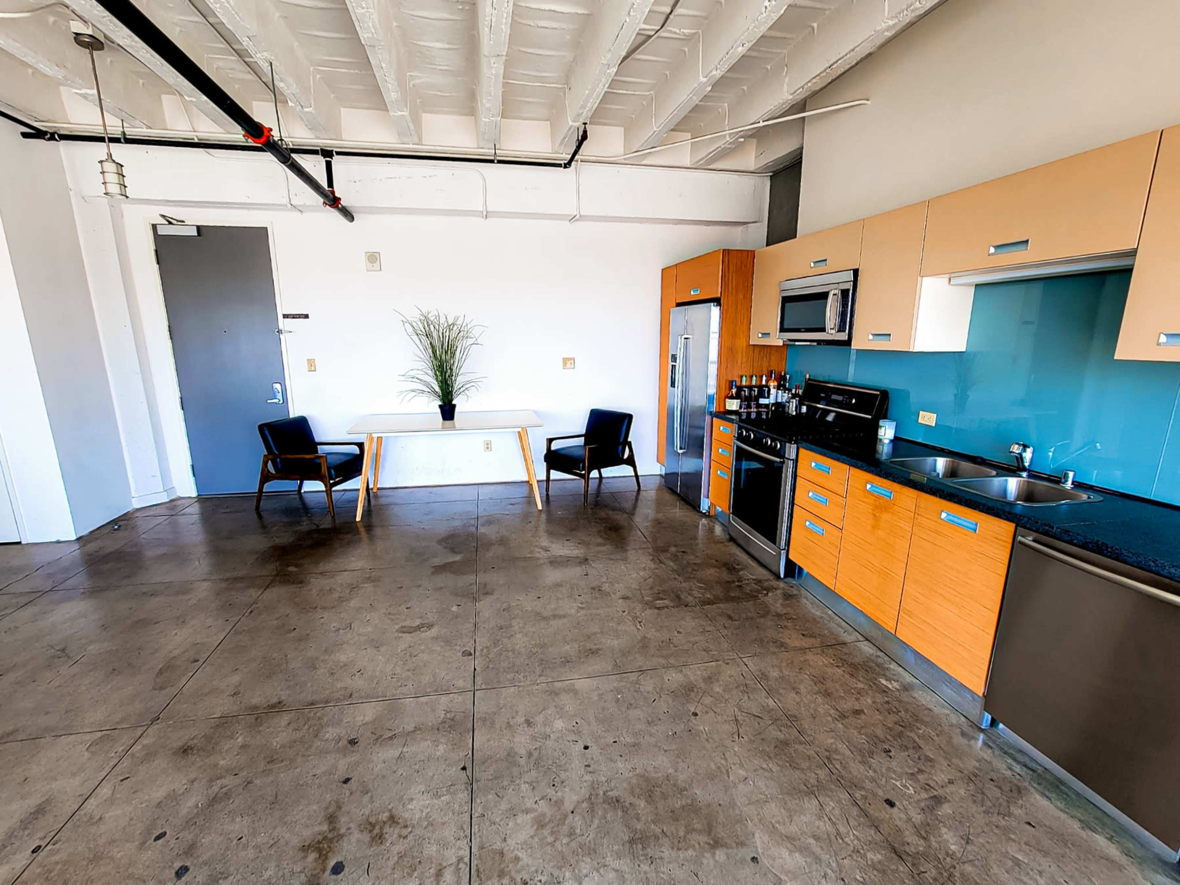 An industrial-style kitchen and dining area features modern wood cabinetry, stainless steel appliances, and a simple table with chairs against a concrete floor.