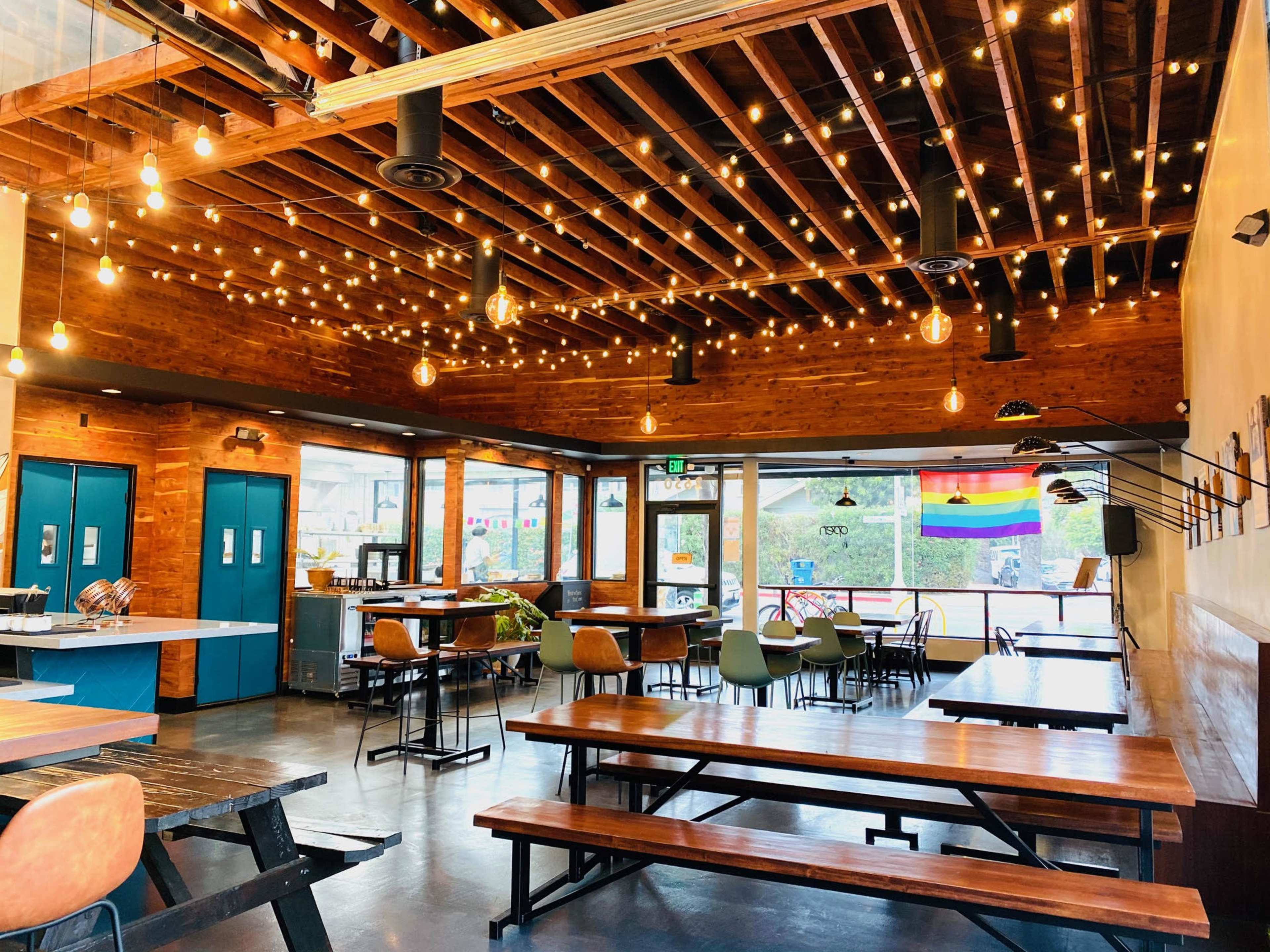 The interior of a restaurant features wooden beams and hanging lights, with tables and chairs arranged throughout the space.