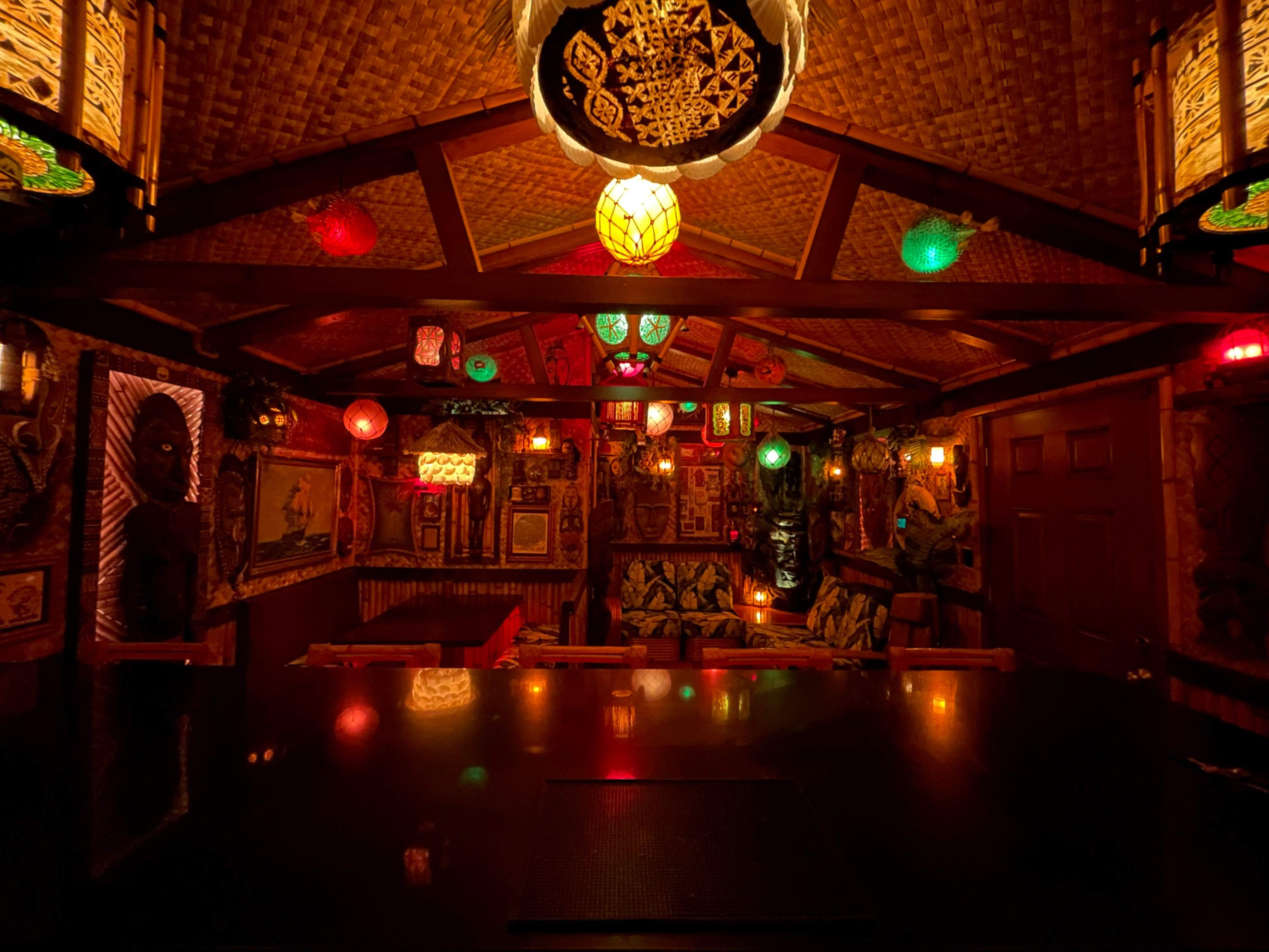 The image depicts a dimly lit tiki bar interior adorned with colorful hanging lanterns and tropical-themed artwork.