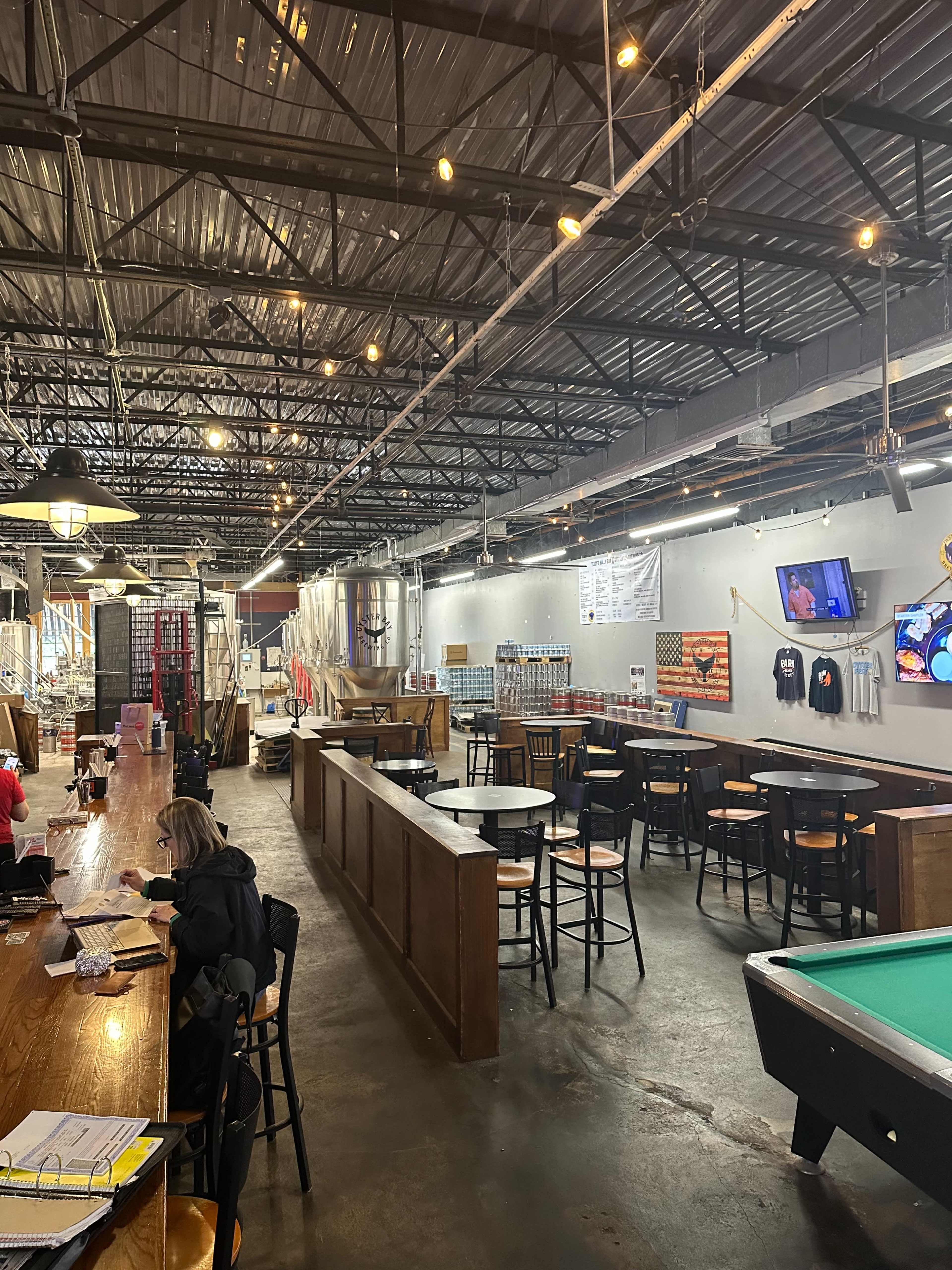 The image shows the interior of a spacious bar or brewery with a long wooden bar, high tables, a pool table, and various brewing equipment in the background.
