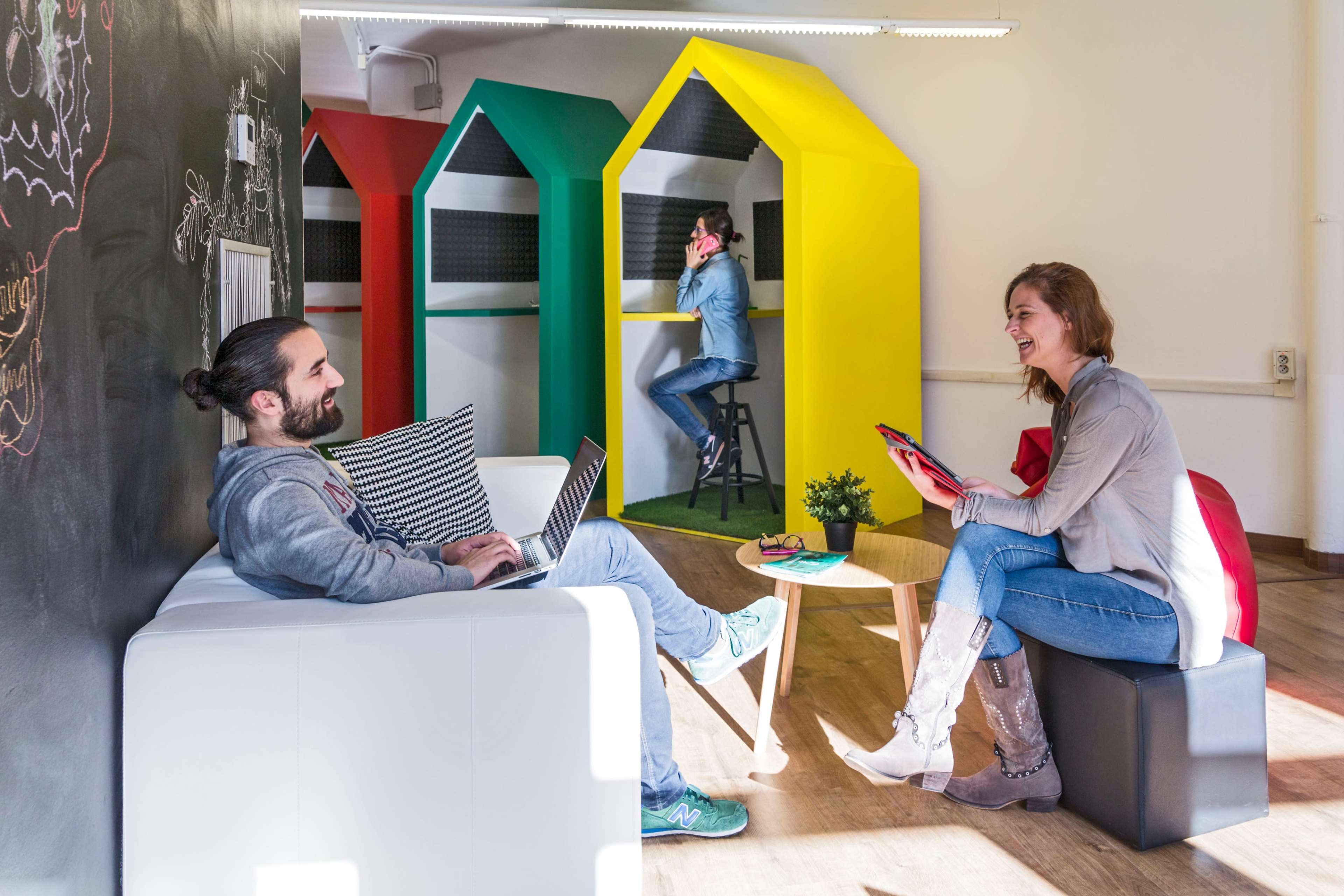 Two people are seated in a bright, modern workspace, one using a laptop on a sofa while the other holds a tablet, with two others in colorful booths in the background.