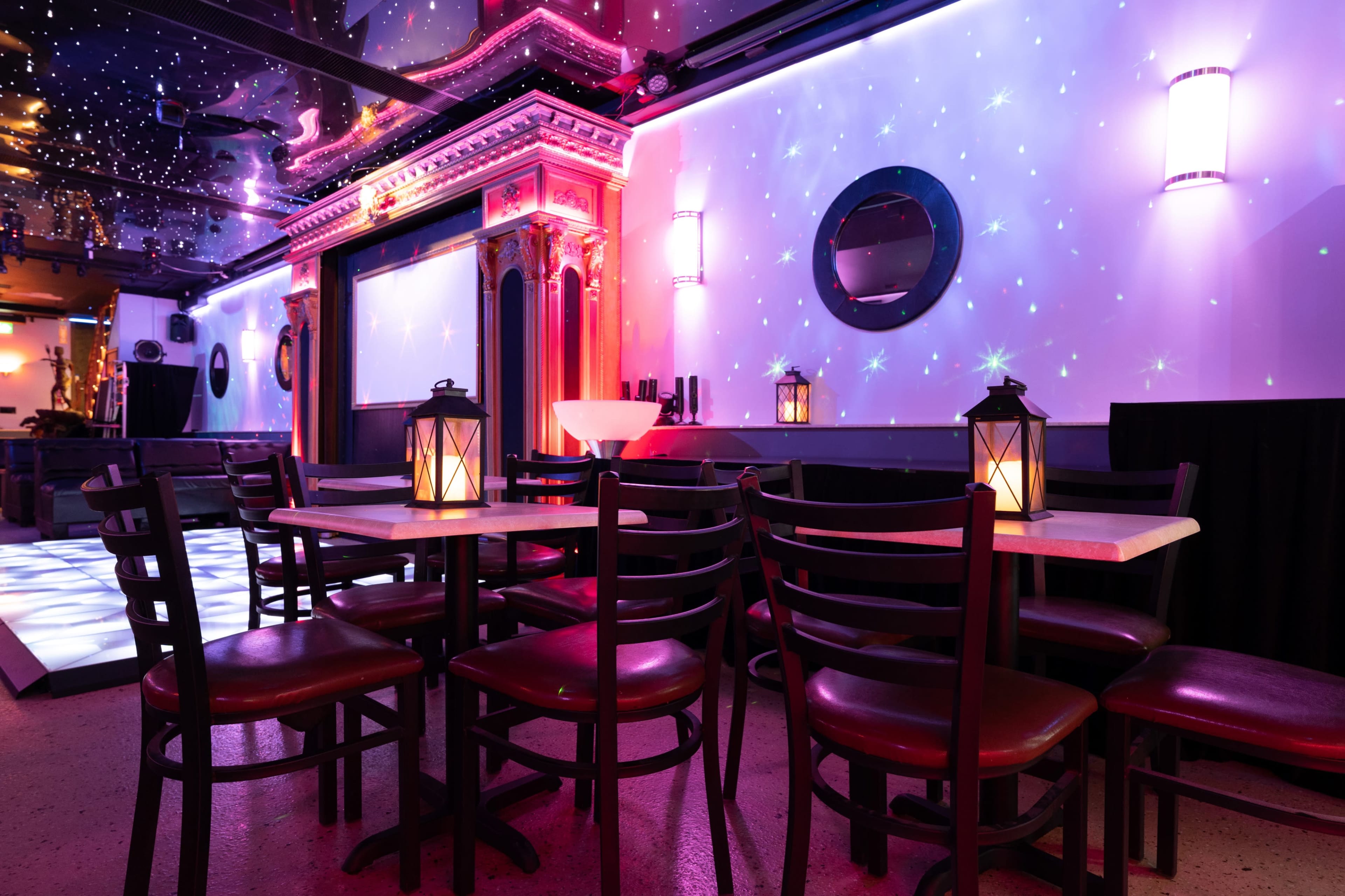 A cozy event space features tables with lanterns, a stage adorned with colorful lighting, and a ceiling resembling a starry night.