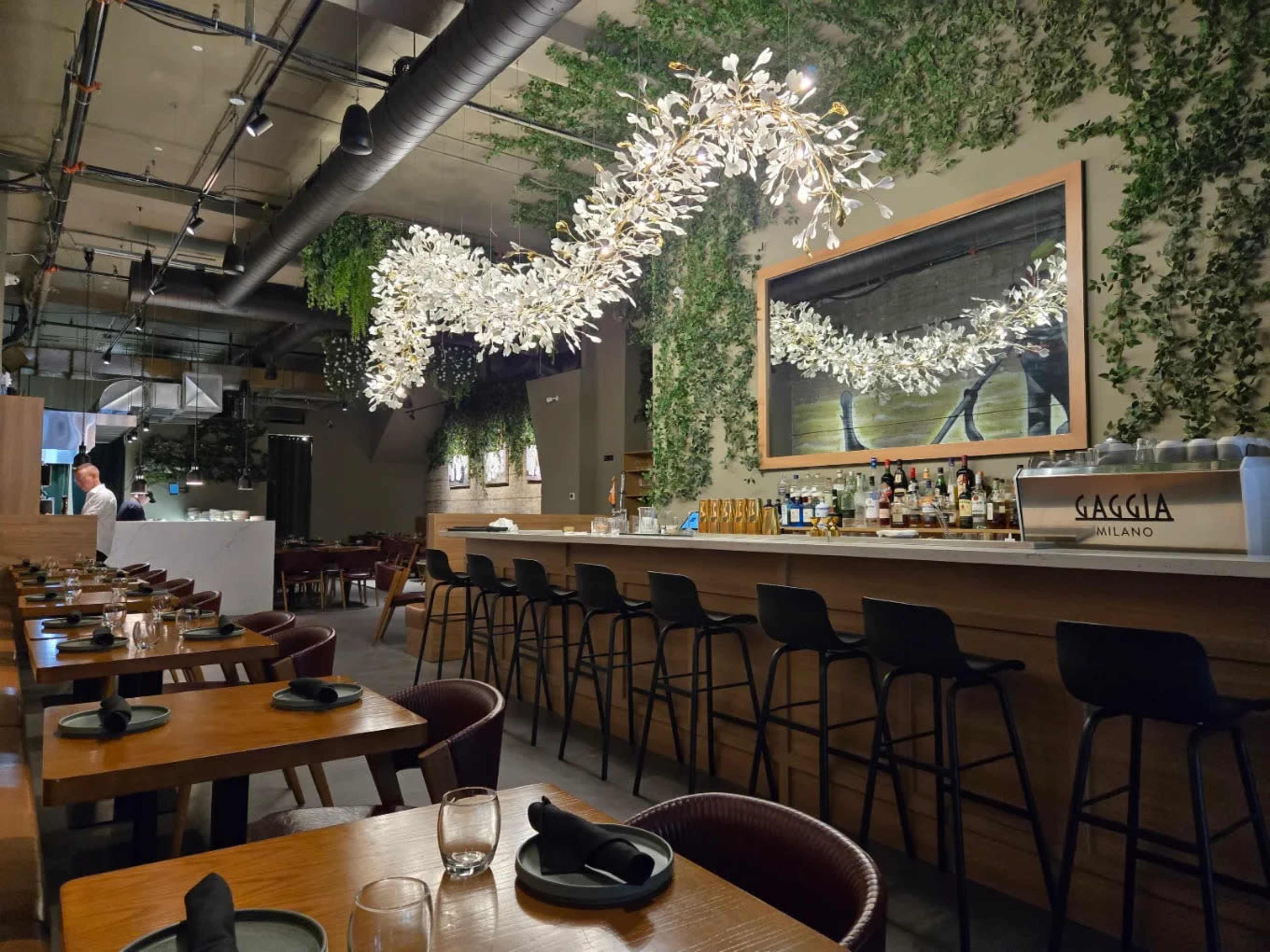 The image shows a modern restaurant interior featuring a bar area with a backlit mirror and an artistic light fixture made of leaves, surrounded by wooden tables and black stools.