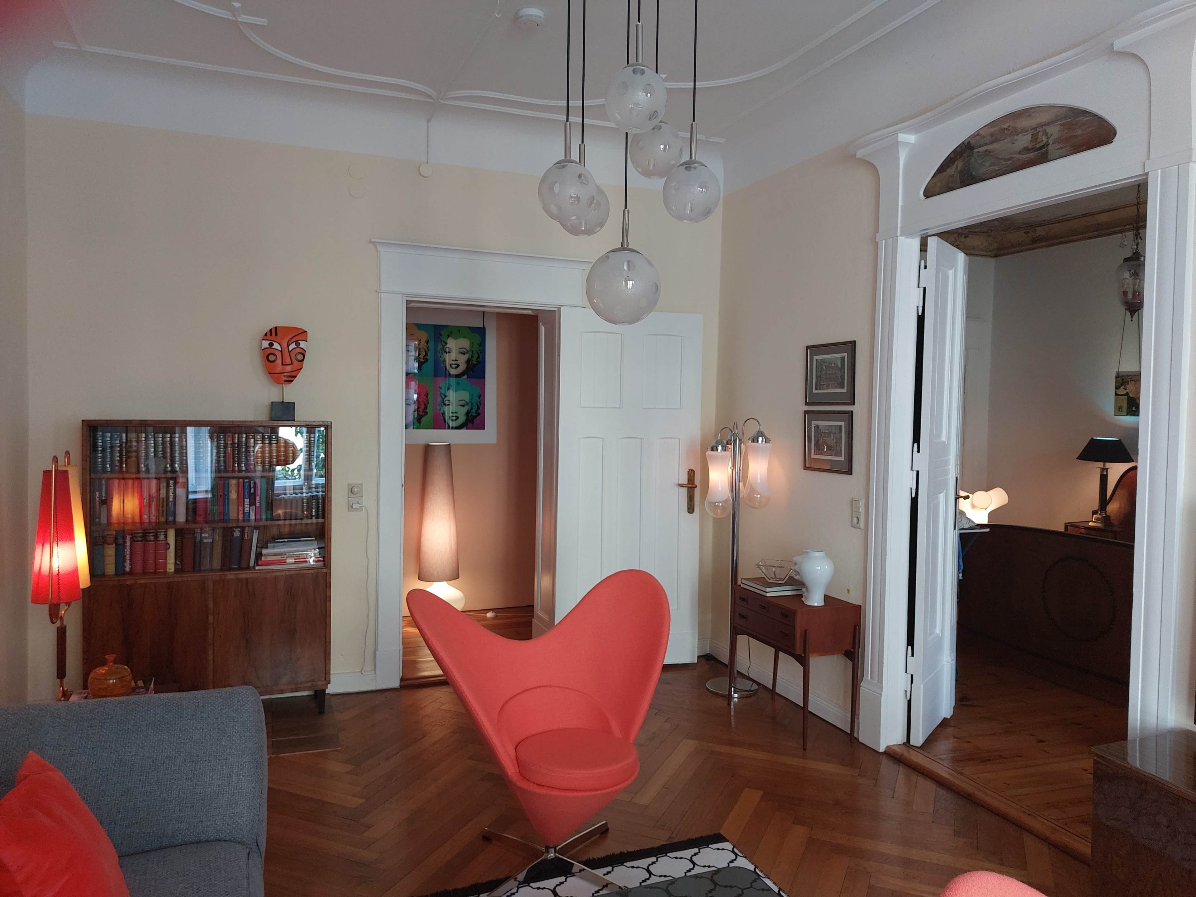 The room features a mid-century modern orange chair, a wooden cabinet with books, and various artwork on the walls, illuminated by pendant lights and lamps.