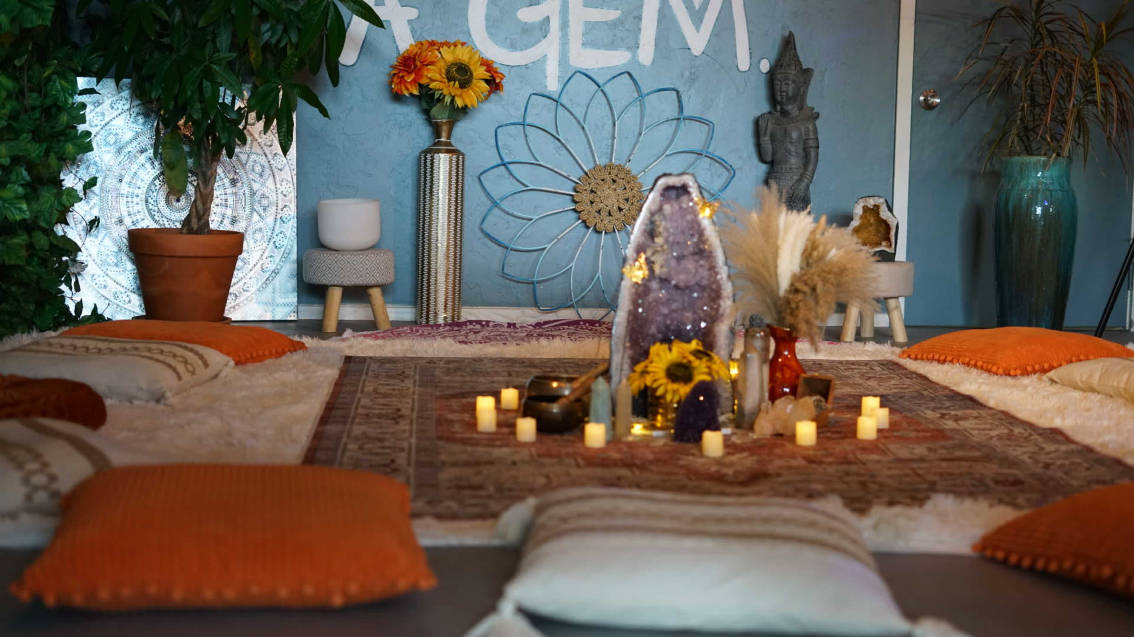 A serene space with cushions surrounding a central arrangement of candles, flowers, and decorative items against a blue wall adorned with a flower design and a statue.