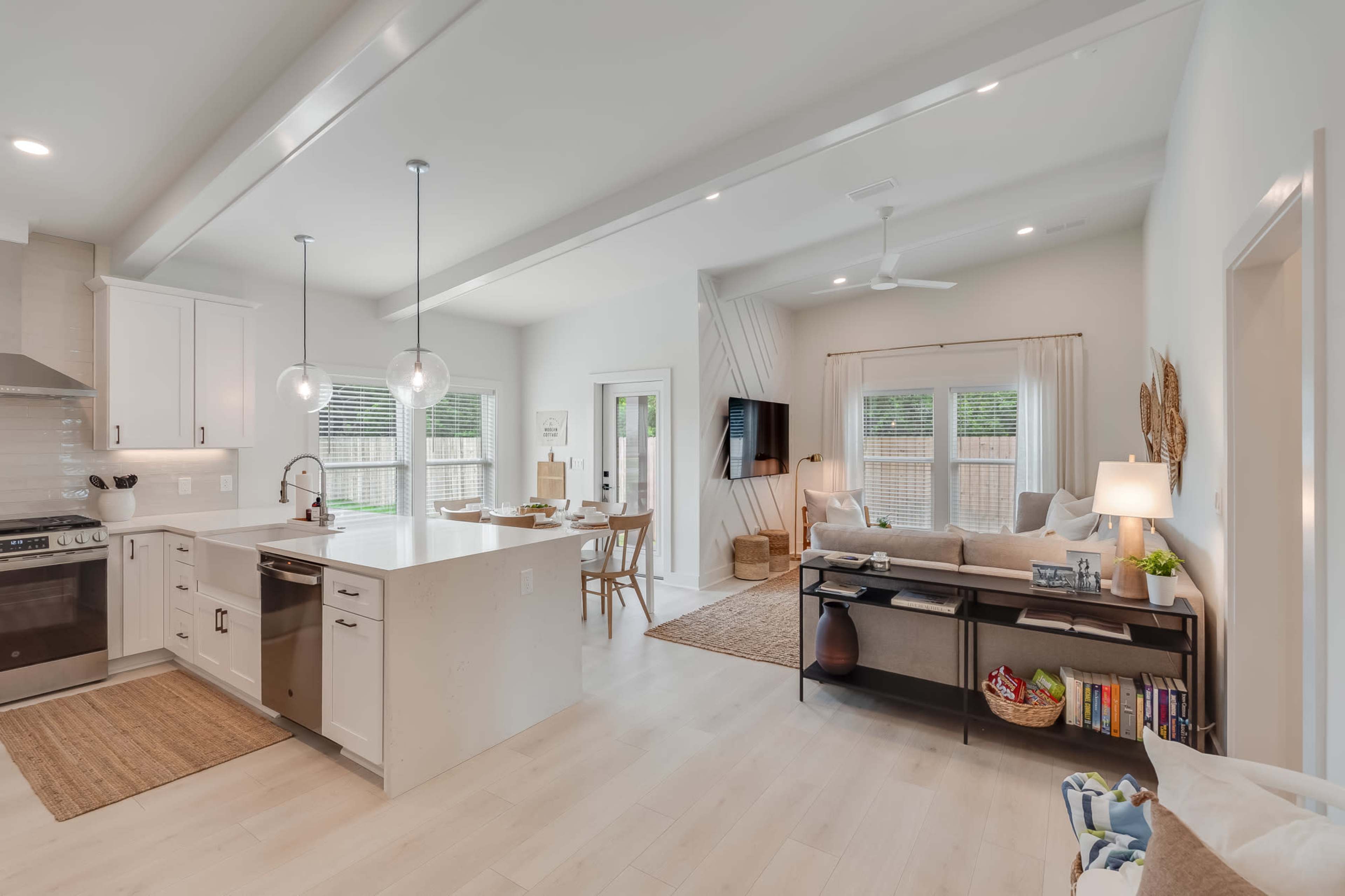 The image shows a modern open-concept kitchen and living area with a light color scheme, featuring a kitchen island, dining table, and seating arrangement.