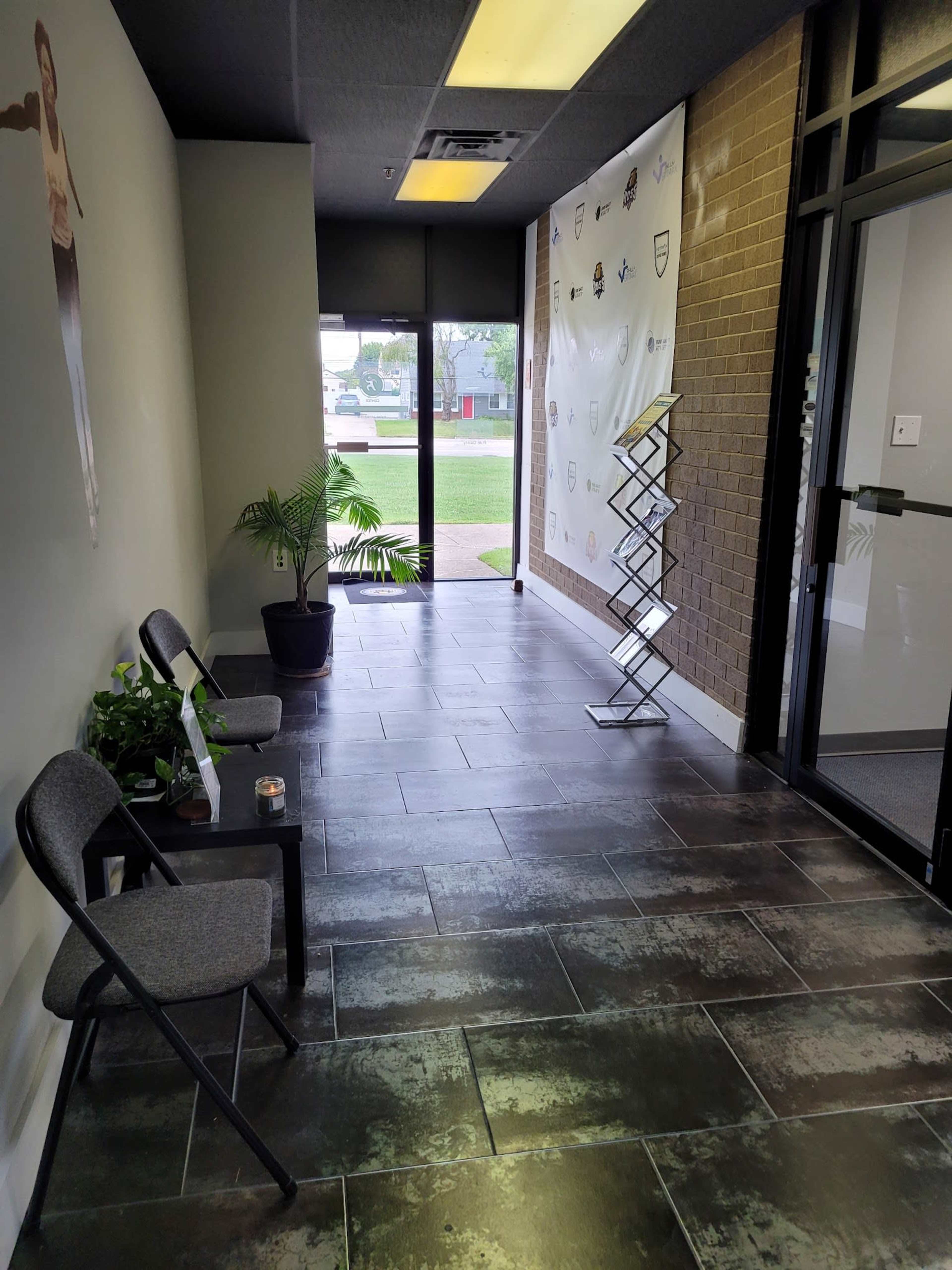 A narrow hallway with chairs, a potted plant, and a wall displaying logos, leading to a glass door with an exterior view.