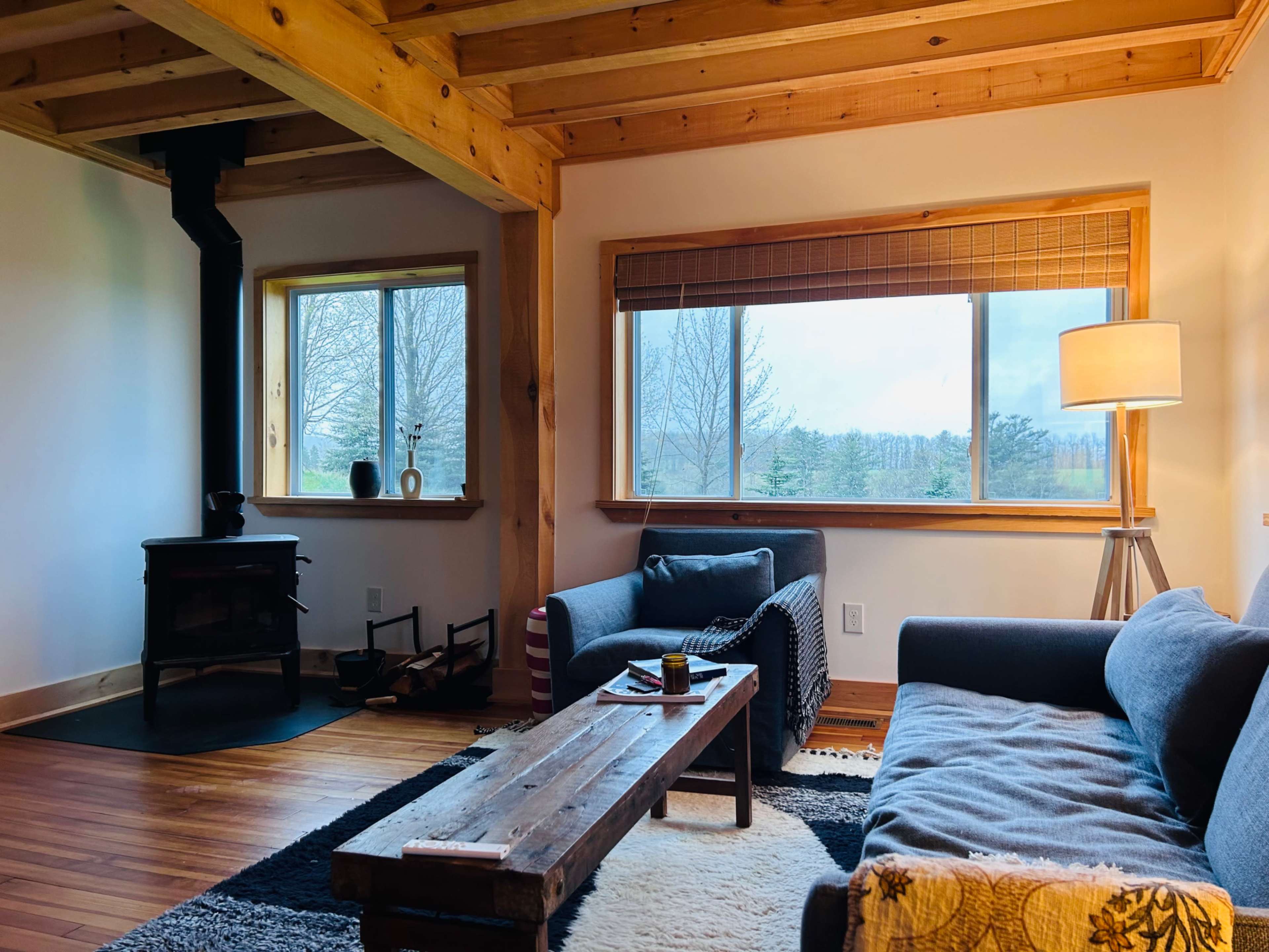A cozy living room with wooden beams, a small stove, two sofas, and a large window overlooking a landscape.