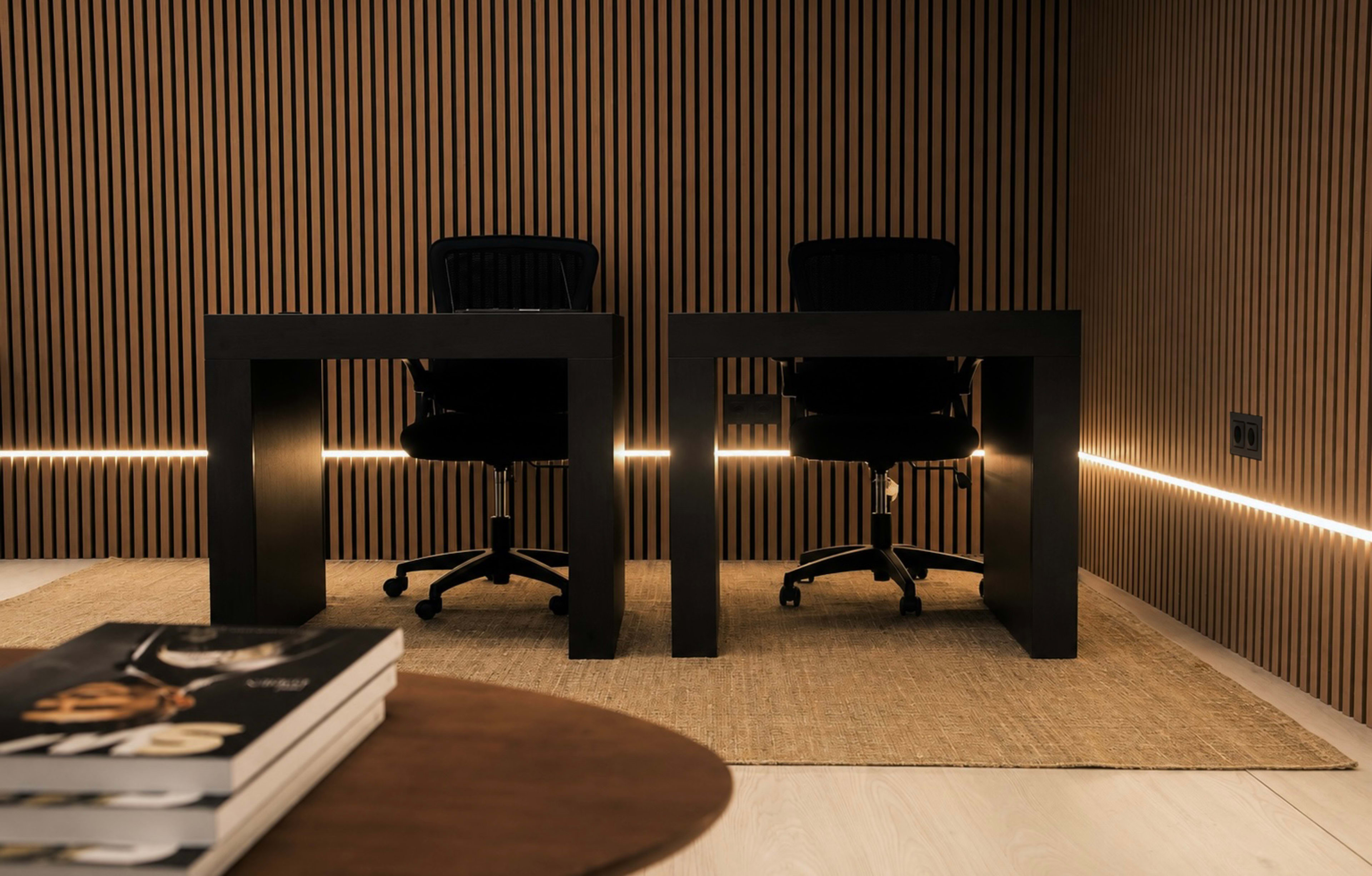 Two black desks with ergonomic chairs are set against a wall featuring vertical wooden slats, with a low table and stacked books in the foreground.