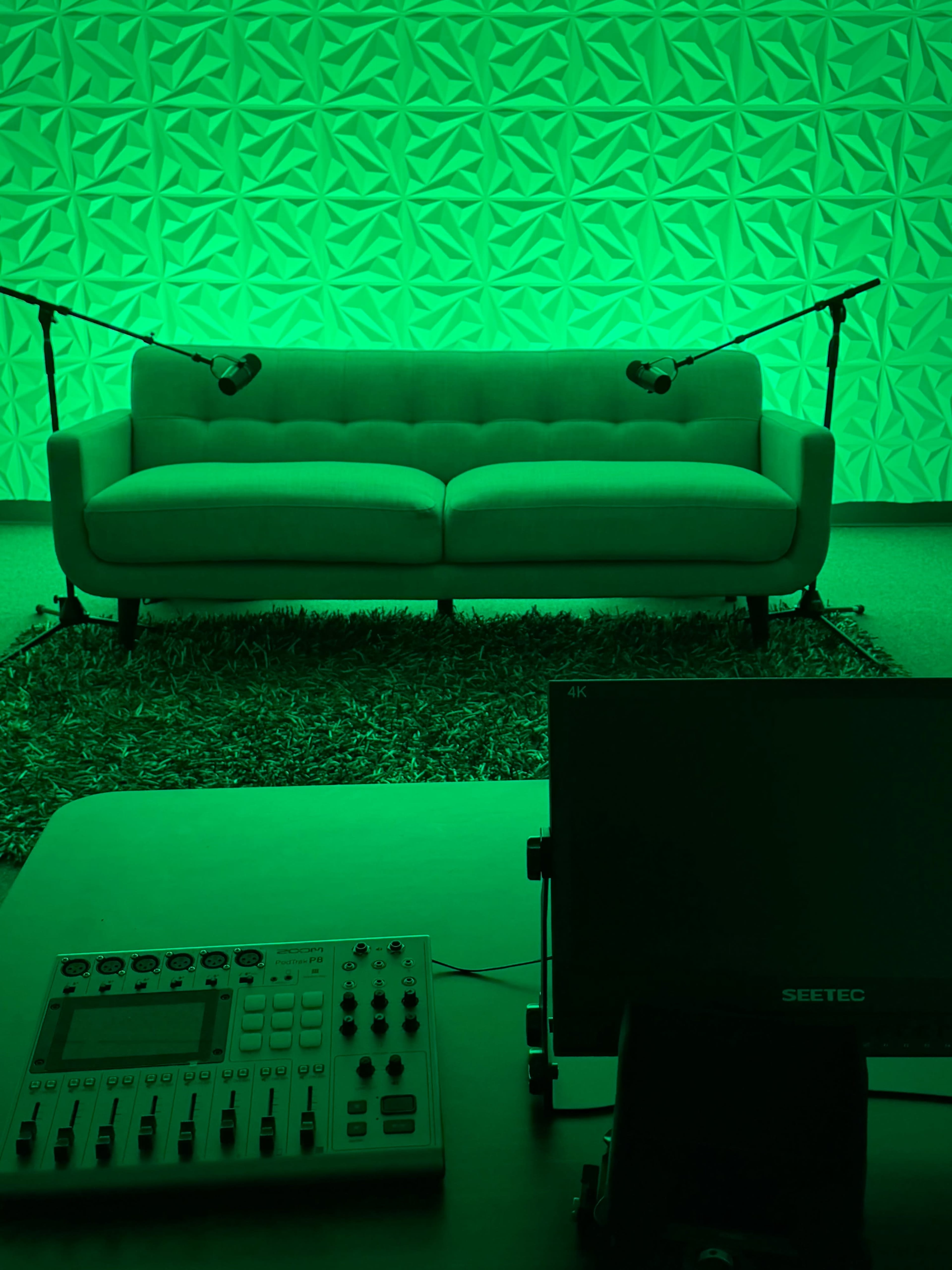 The image shows a green-lit recording studio with a couch, two microphones, and audio equipment on a table.