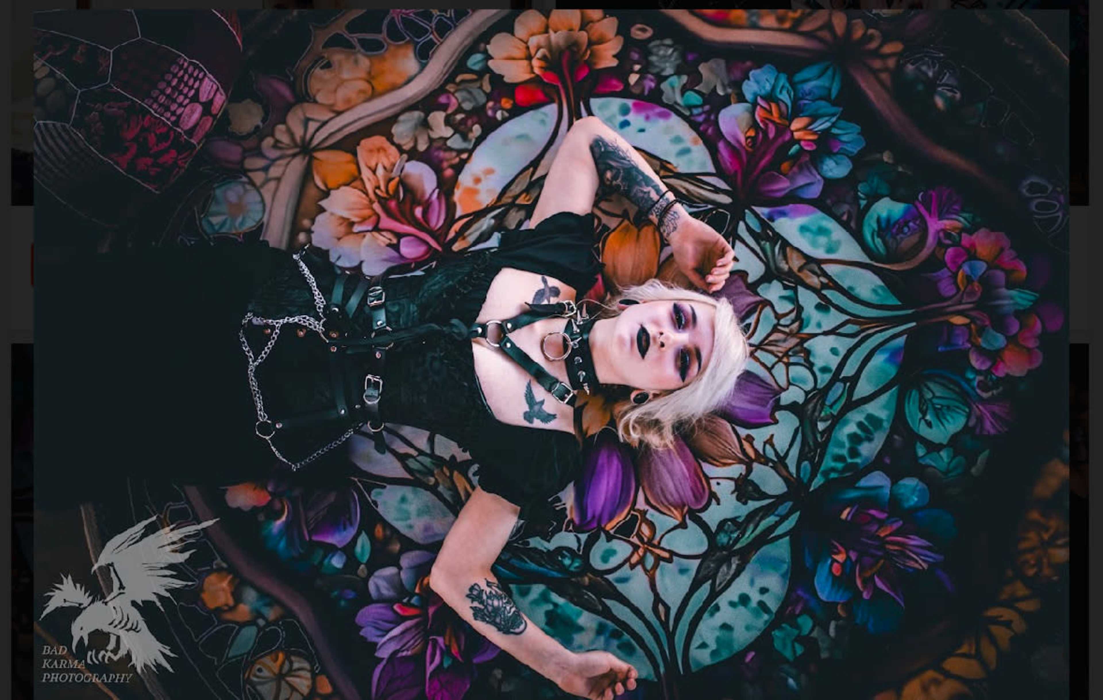 A person lies on a colorful floral-patterned rug, surrounded by vibrant artwork, wearing a dark outfit with chains and tattoos visible on their arms.