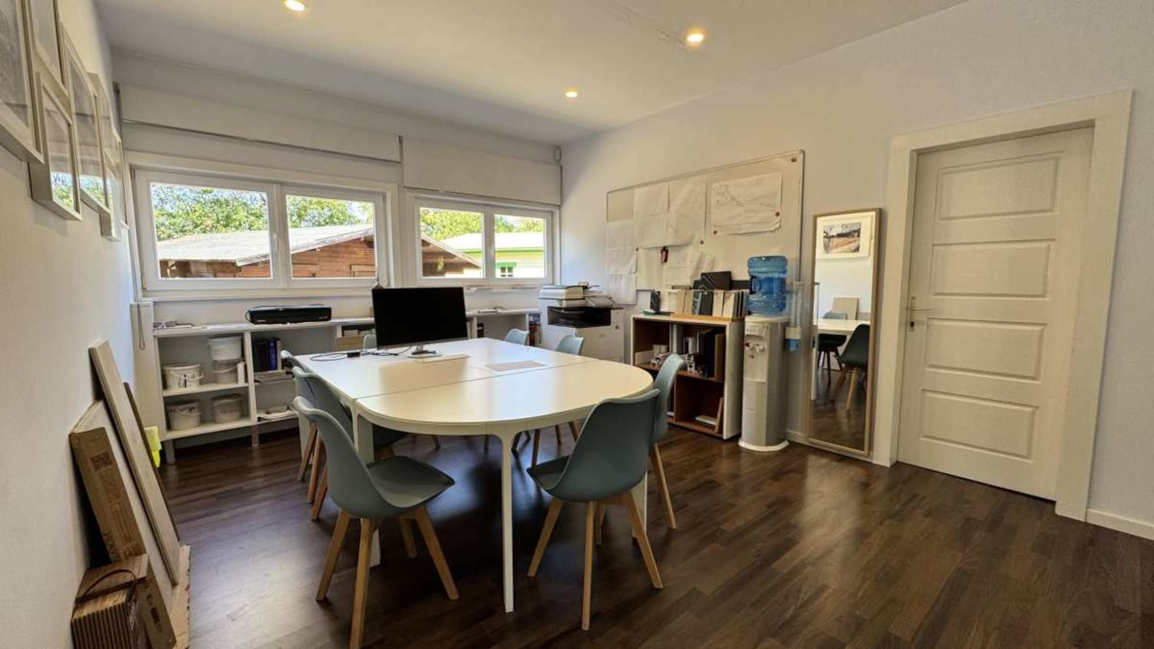 A bright and organized office space features a white table surrounded by modern chairs, a water cooler, and large windows.