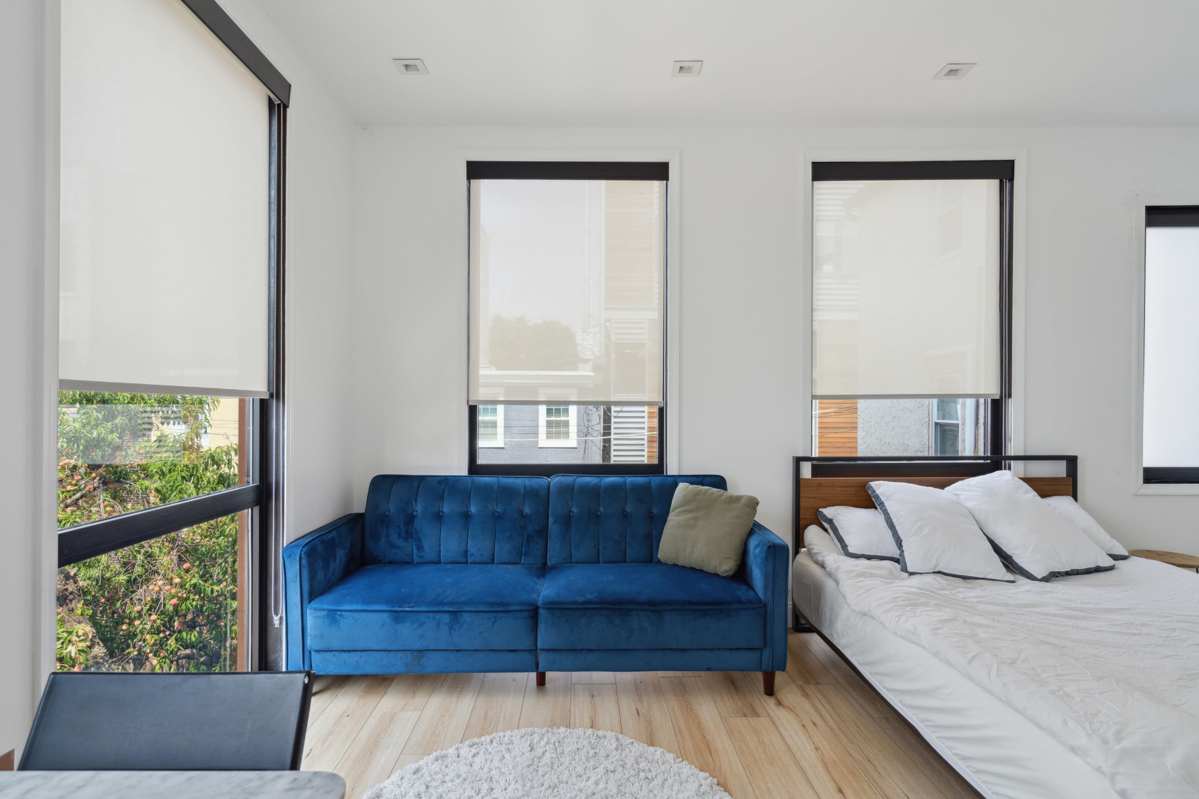 The image shows a bright, minimalist bedroom featuring a blue velvet sofa, a wooden bed with unmade white linens, and large windows with light curtains.