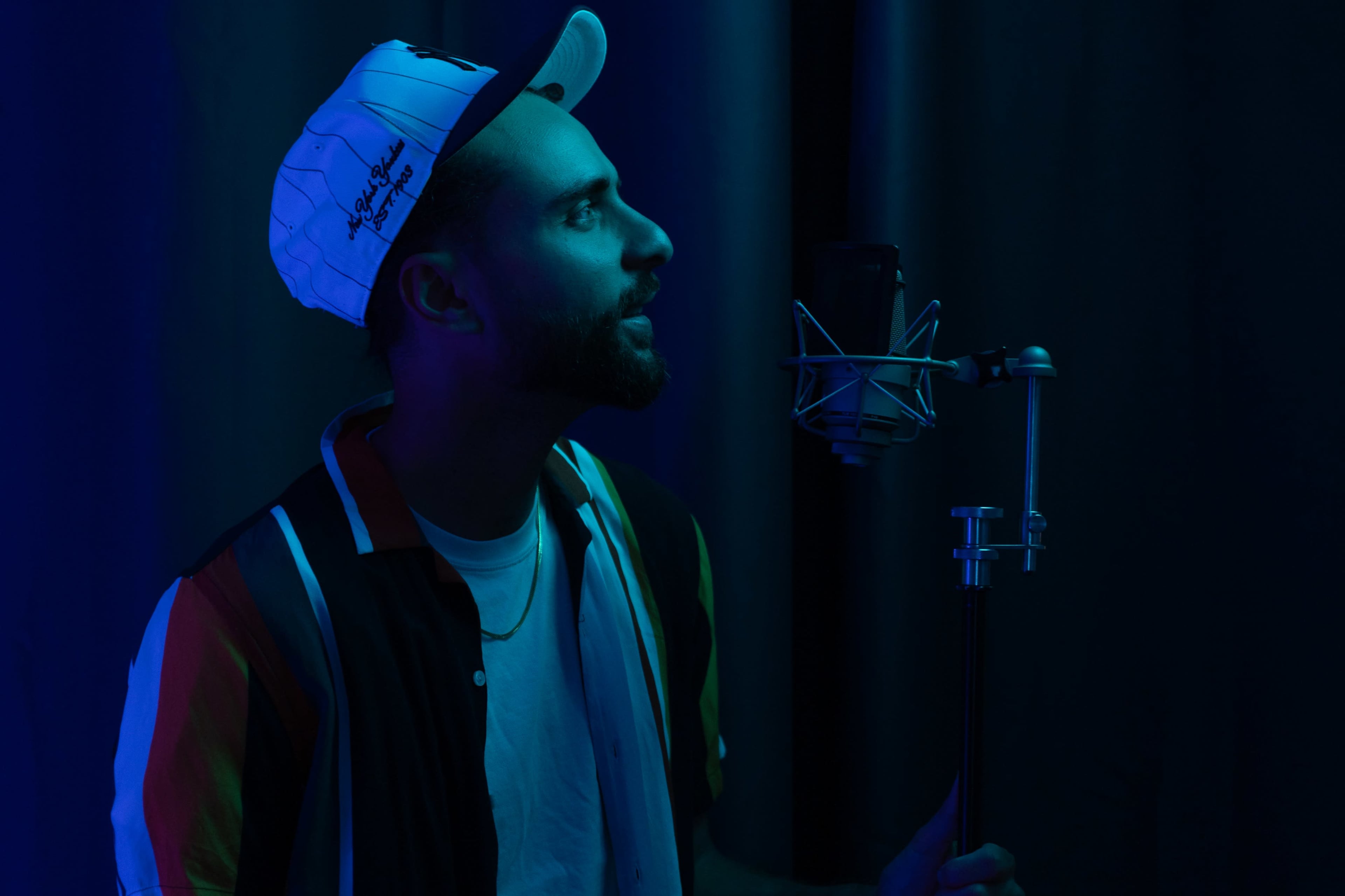 A man with a cap stands next to a microphone in a dimly lit room with blue lighting.