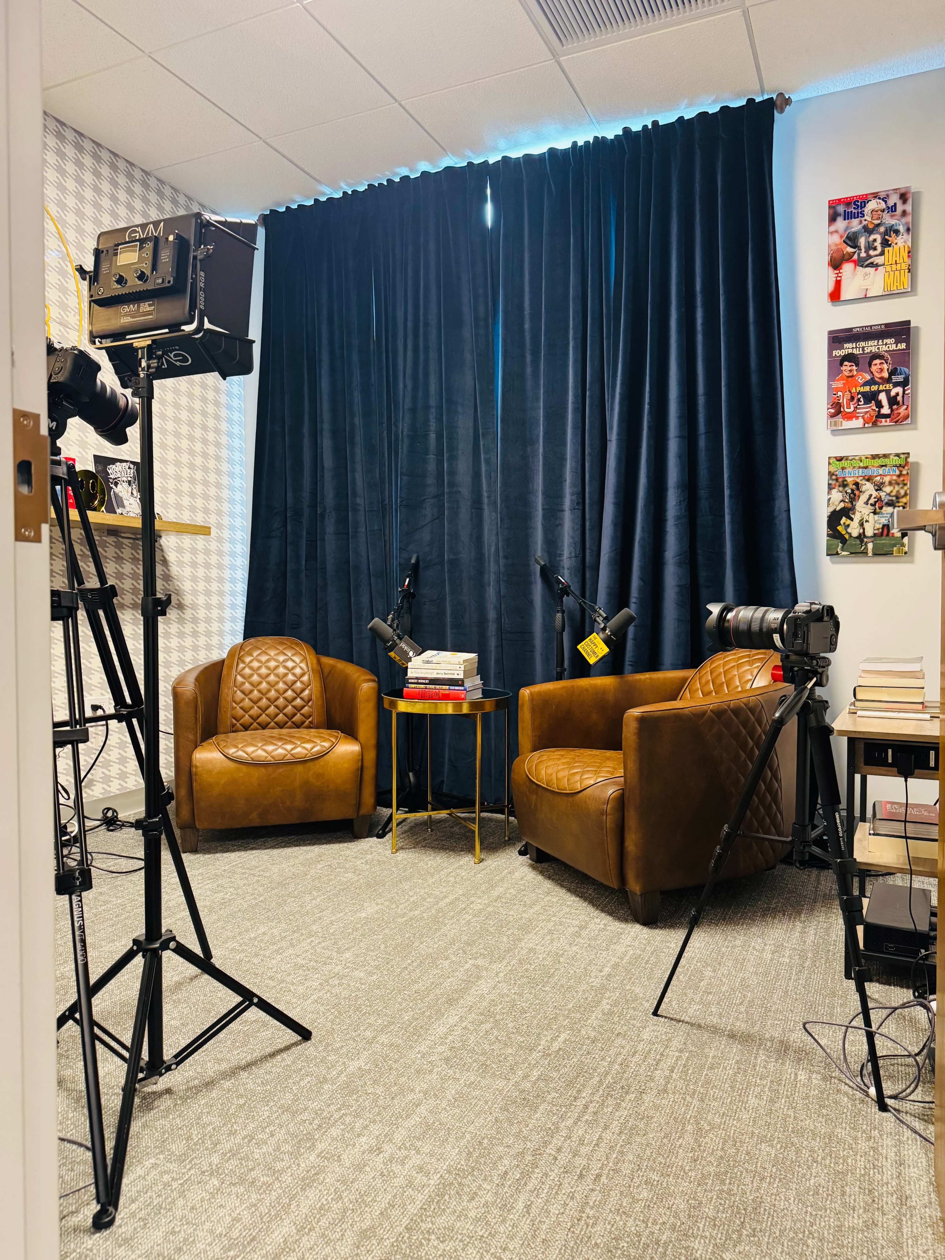 The image shows a small recording studio featuring two brown leather chairs, a table with books, and cameras on tripods, all set against a backdrop of dark curtains and framed pictures on the walls.
