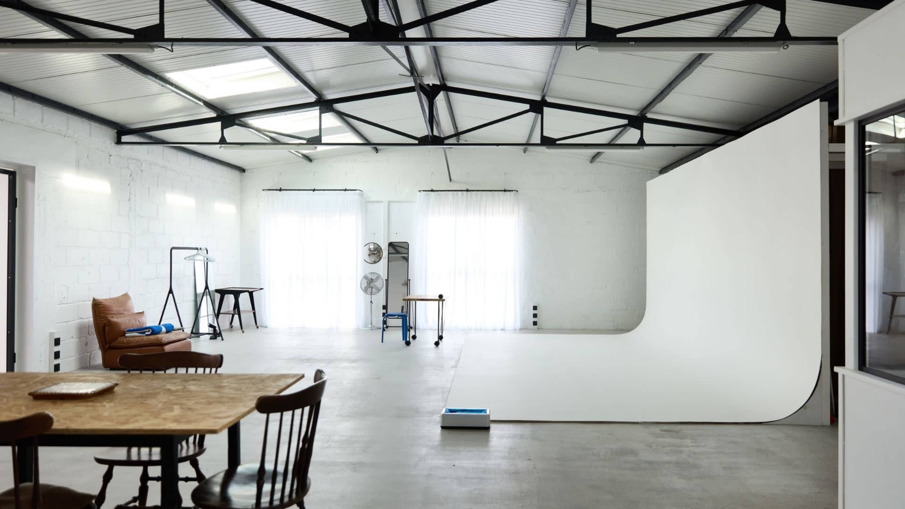 The image shows a spacious photography studio with a white backdrop, natural light streaming through large windows, and a wooden table with chairs.