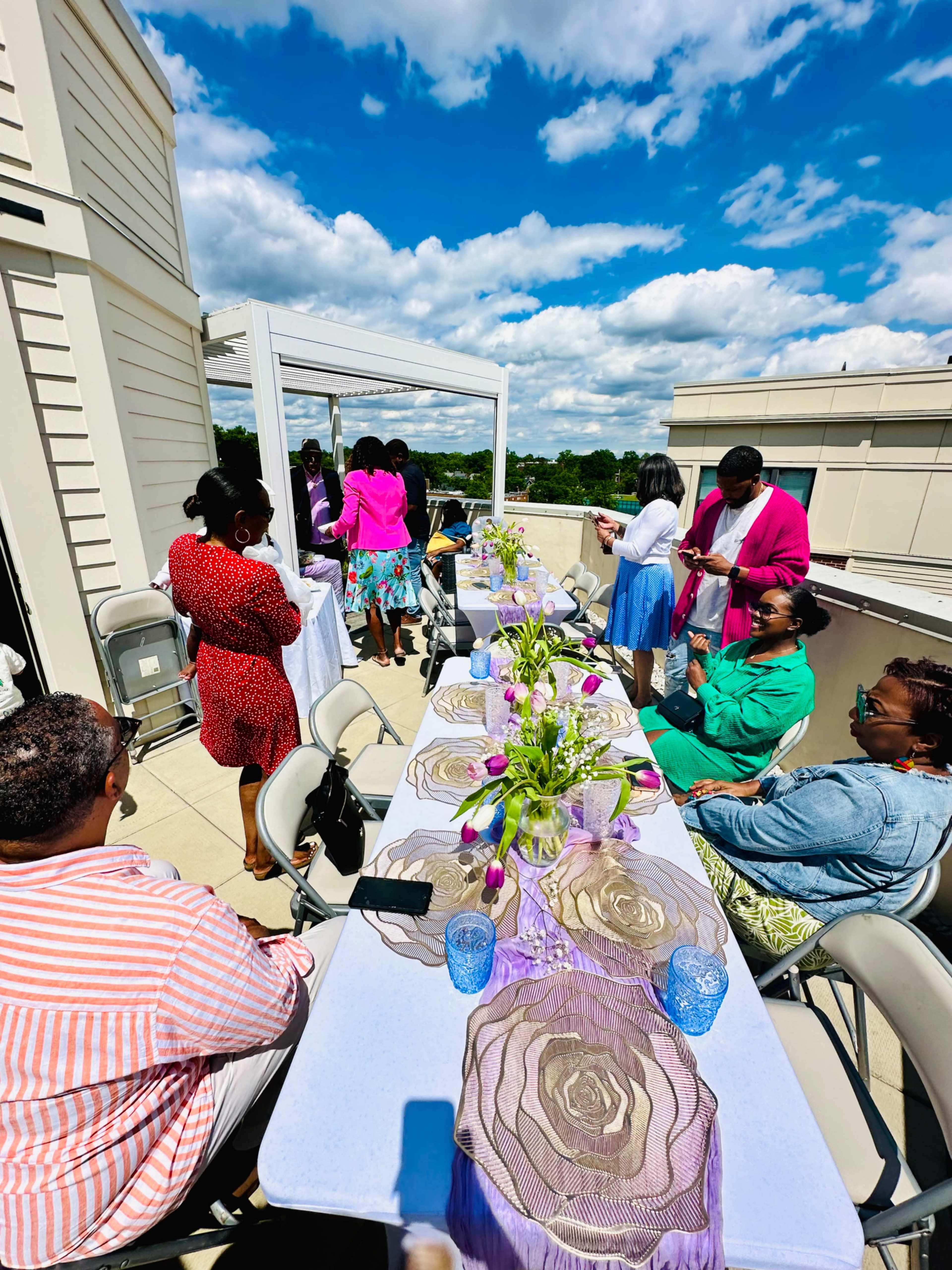 A group of people gather around decorated tables on a rooftop, enjoying a sunny day with flowers and glassware.