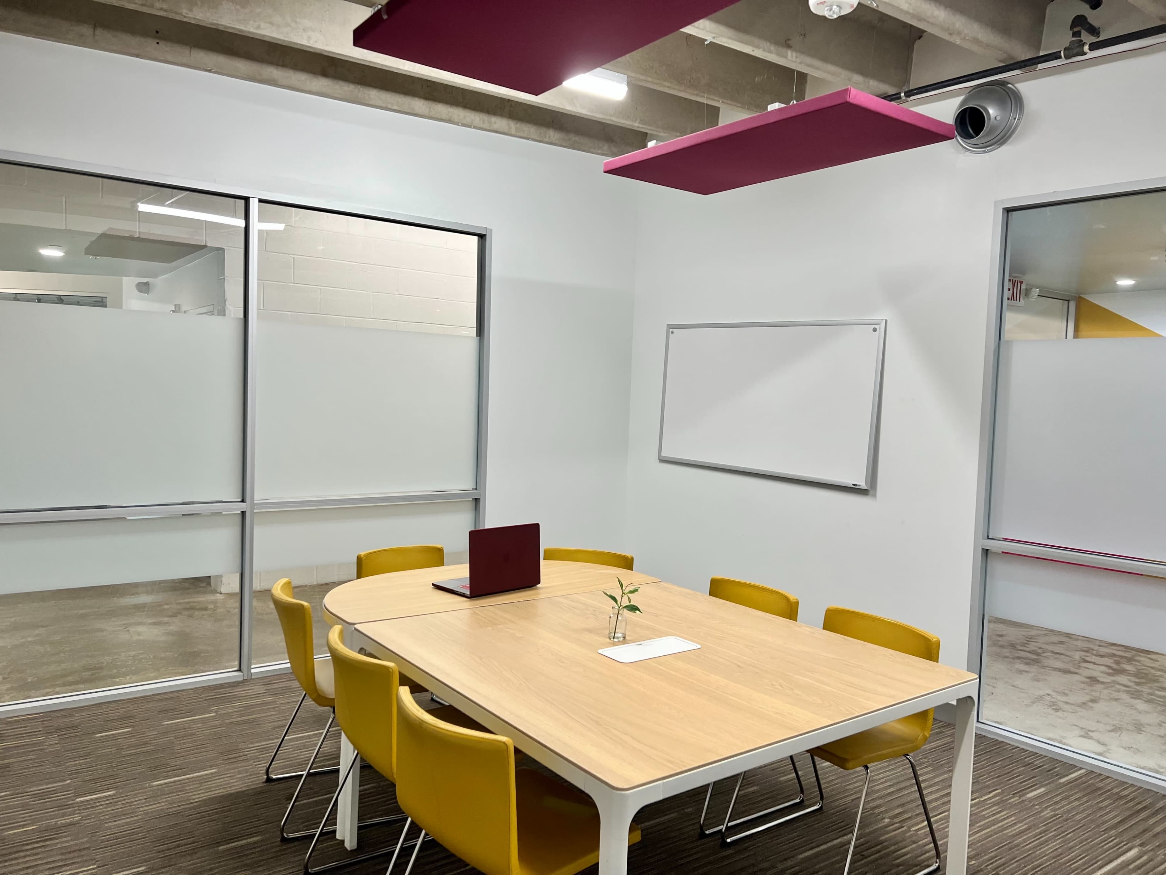 A meeting room features a wooden table surrounded by yellow chairs, with a laptop on the table and a small plant in the center.
