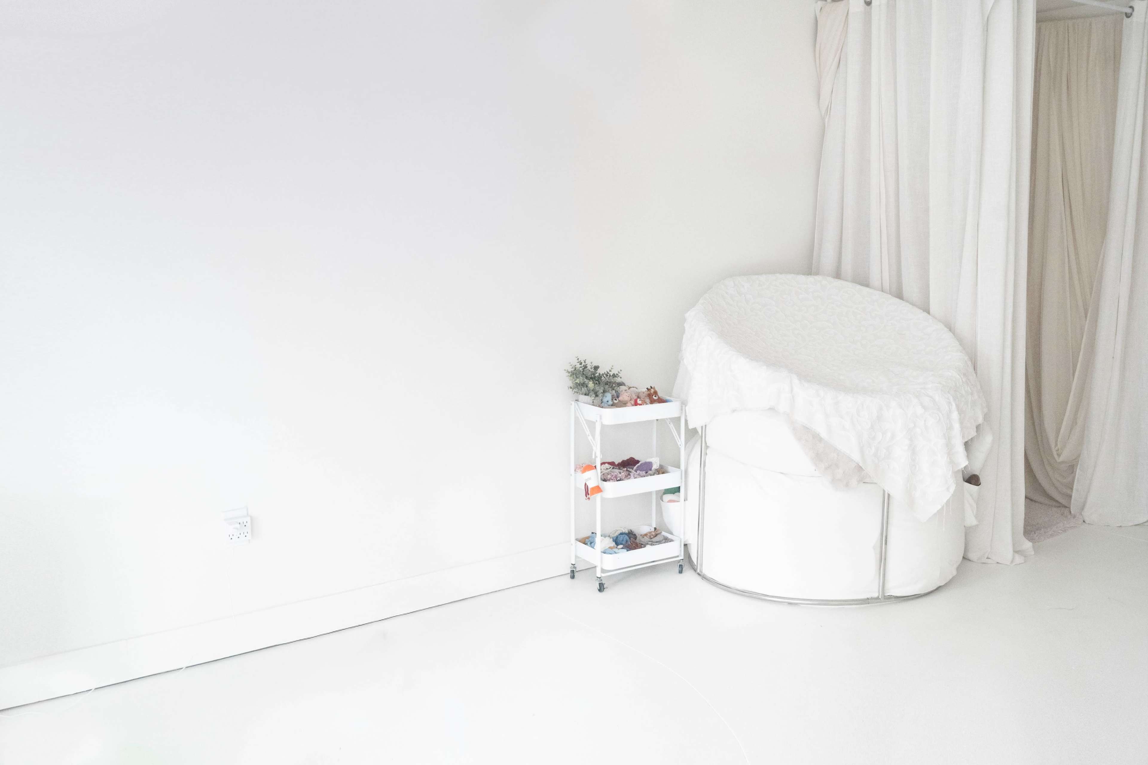 The image shows a minimalist room with a white circular chair draped in a fluffy cover and a small, wheeled cart containing various items.