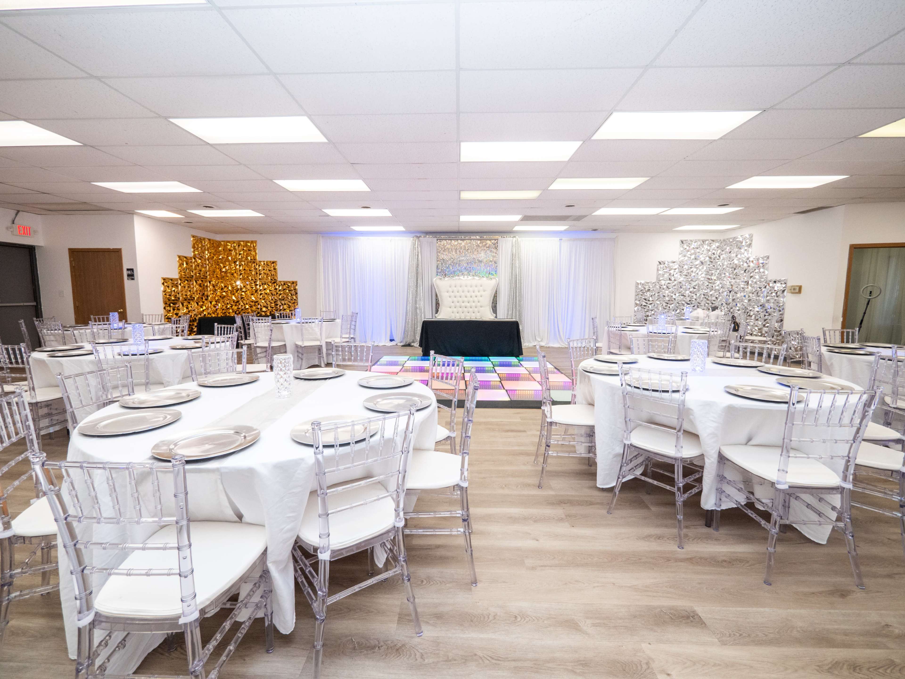 A banquet hall set up with round tables and clear chiavari chairs, featuring decorative backdrops on the walls and a central stage area.