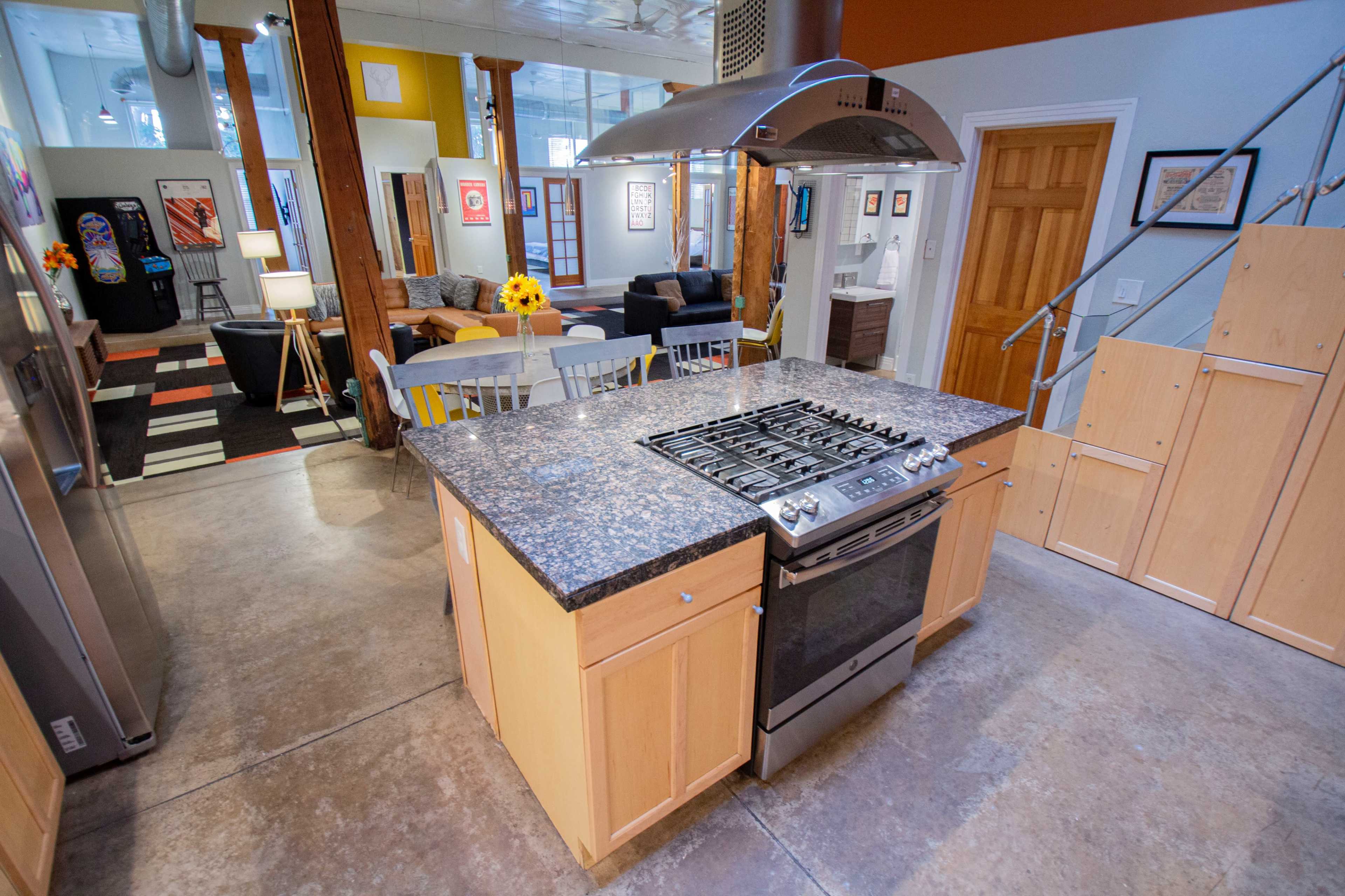 The image shows a modern kitchen with a central island, gas stove, and stainless steel appliances, leading into a living area with wooden beams and a separated seating space.