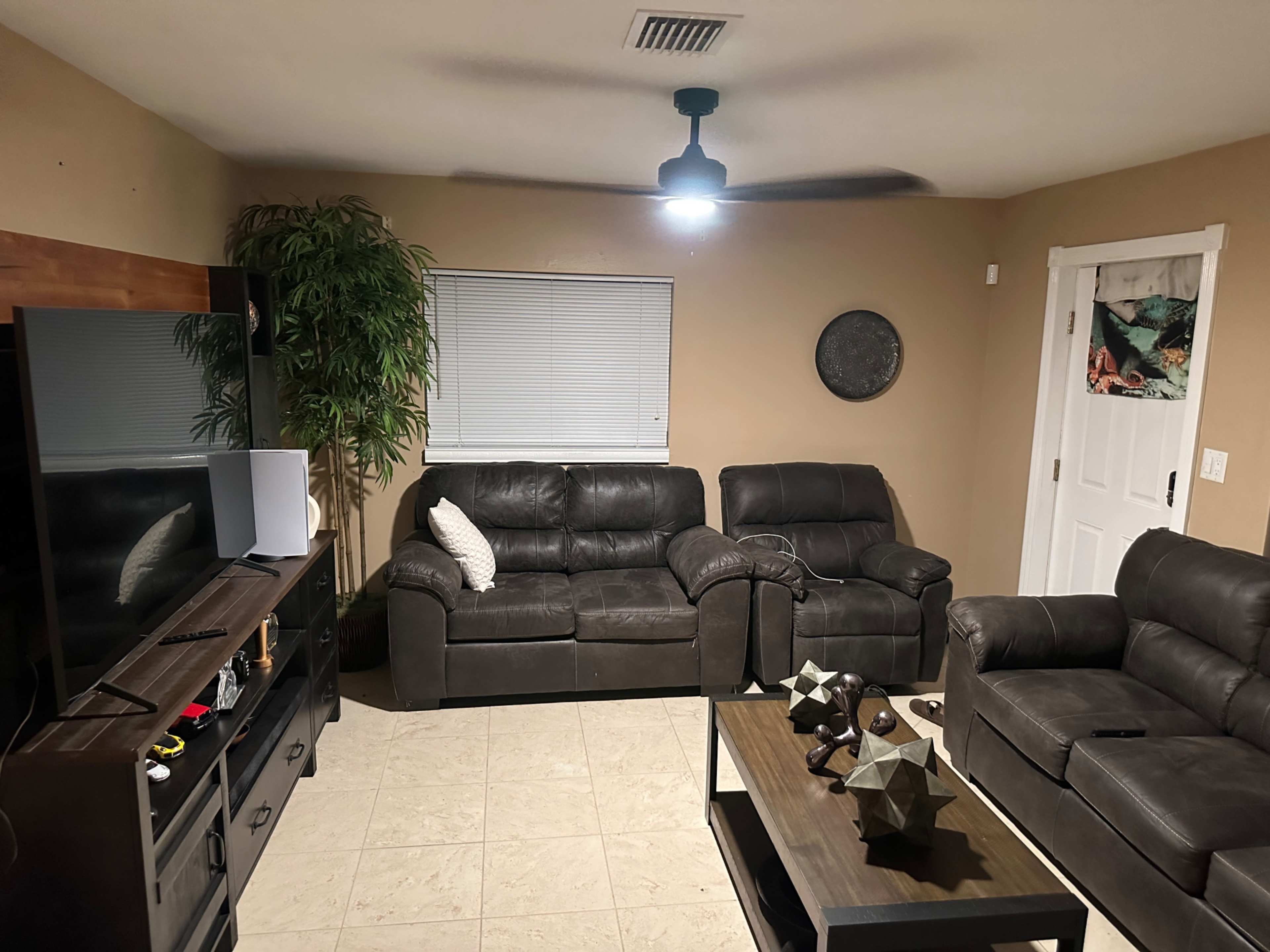 A living room with two dark leather couches, a coffee table with decorative objects, a television, and a door, all set against beige walls and tile flooring.