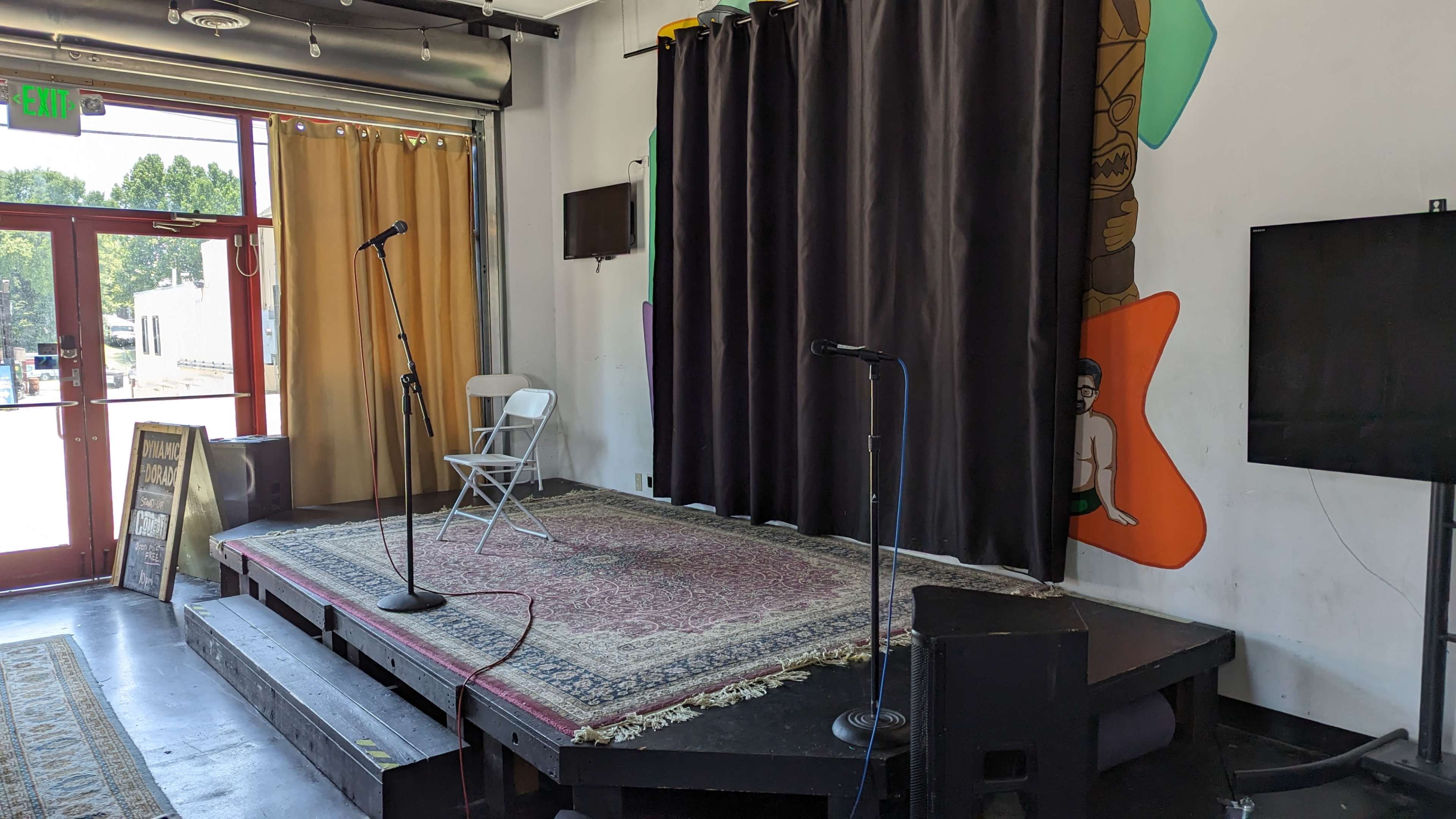 A small stage with two microphones, a white chair, and a patterned rug, set against a backdrop of curtains and artwork on the wall.