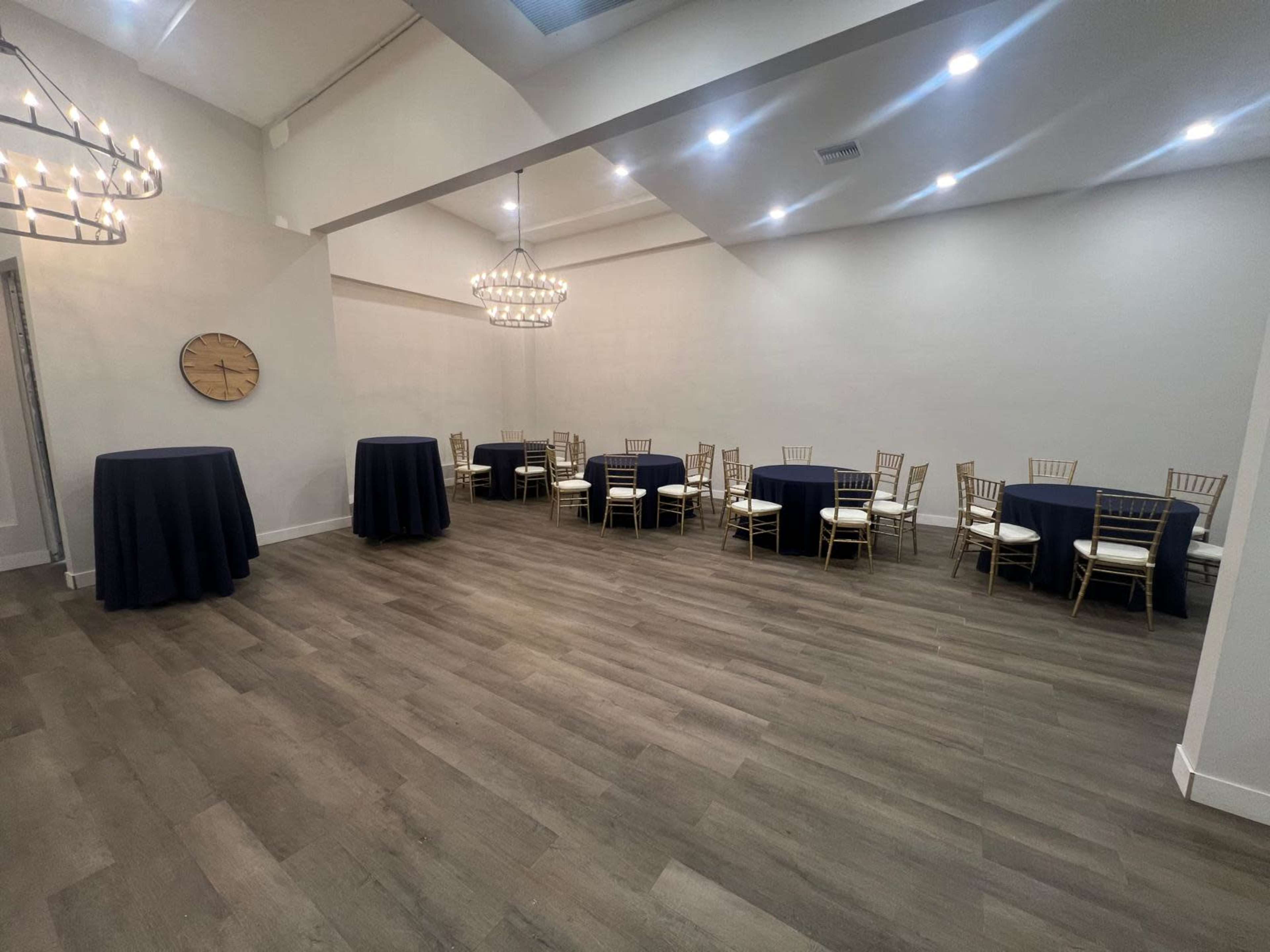 The image shows a spacious event room set with round tables and tall cocktail tables, illuminated by hanging light fixtures.