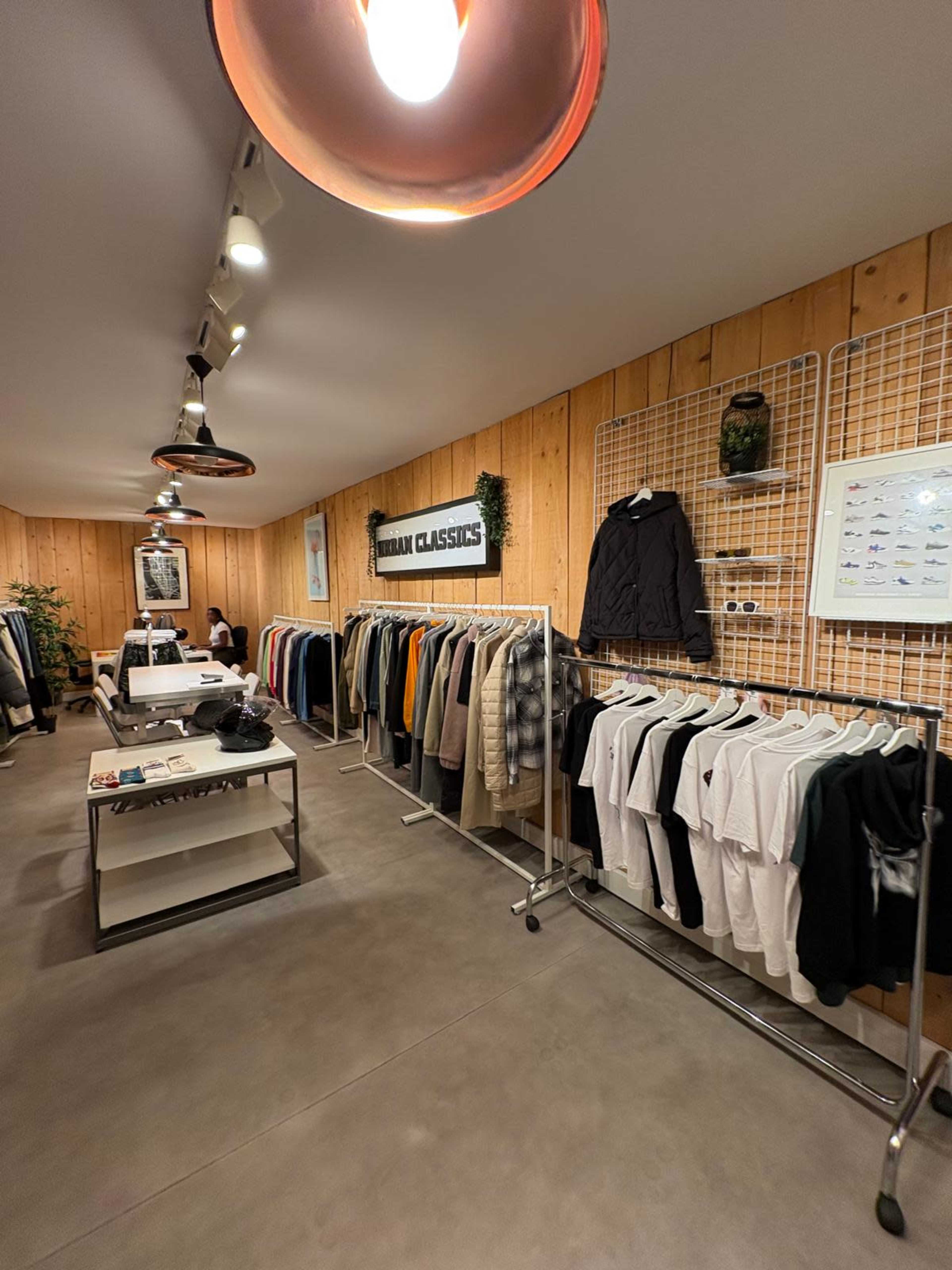 The image shows a modern retail space with clothing displays along the walls, a central table with various items, and a welcoming atmosphere created by wooden paneling and overhead lighting.