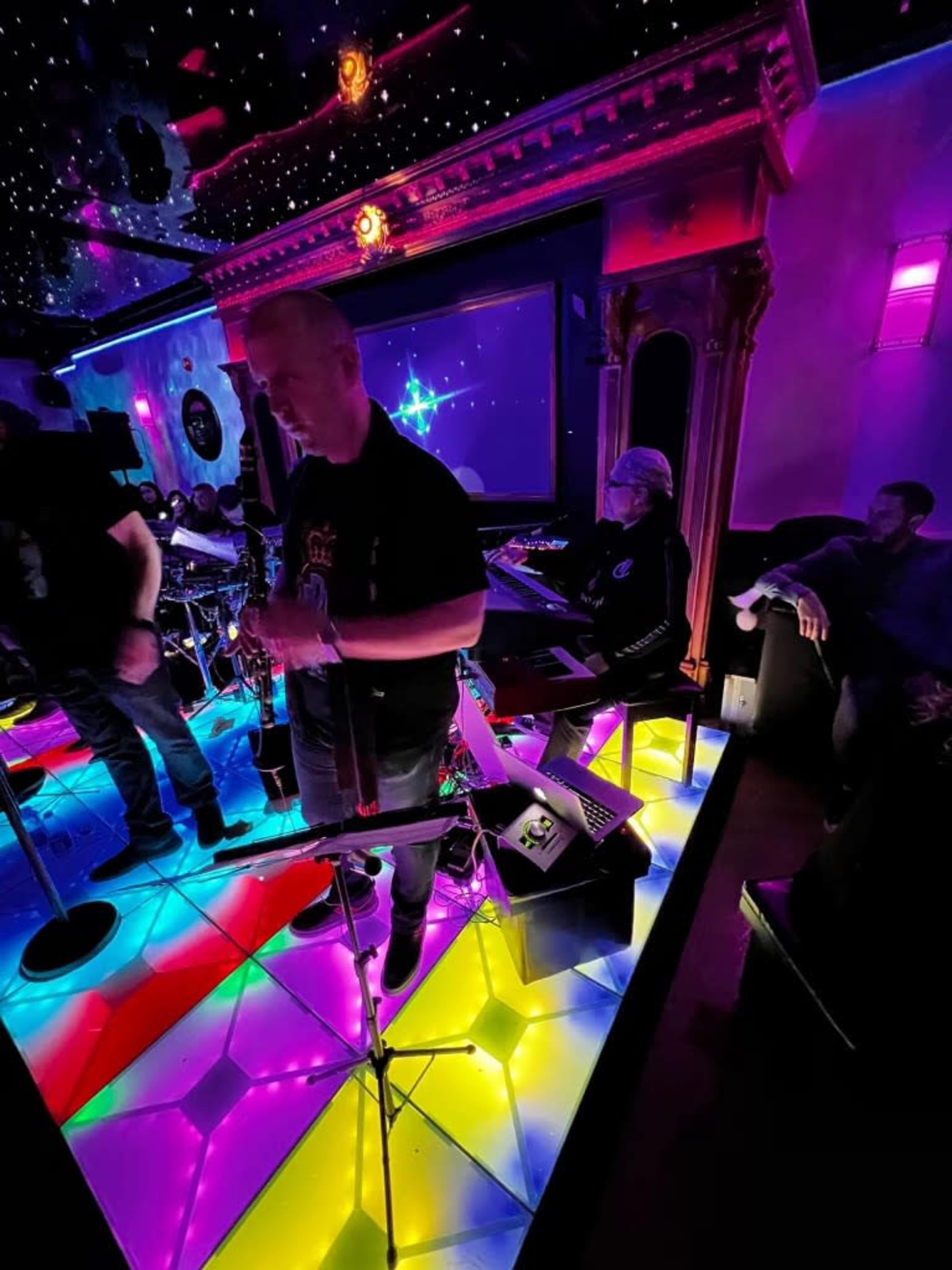 A brightly lit bar or club features a colorful floor with musicians performing on stage and seated patrons in the background.