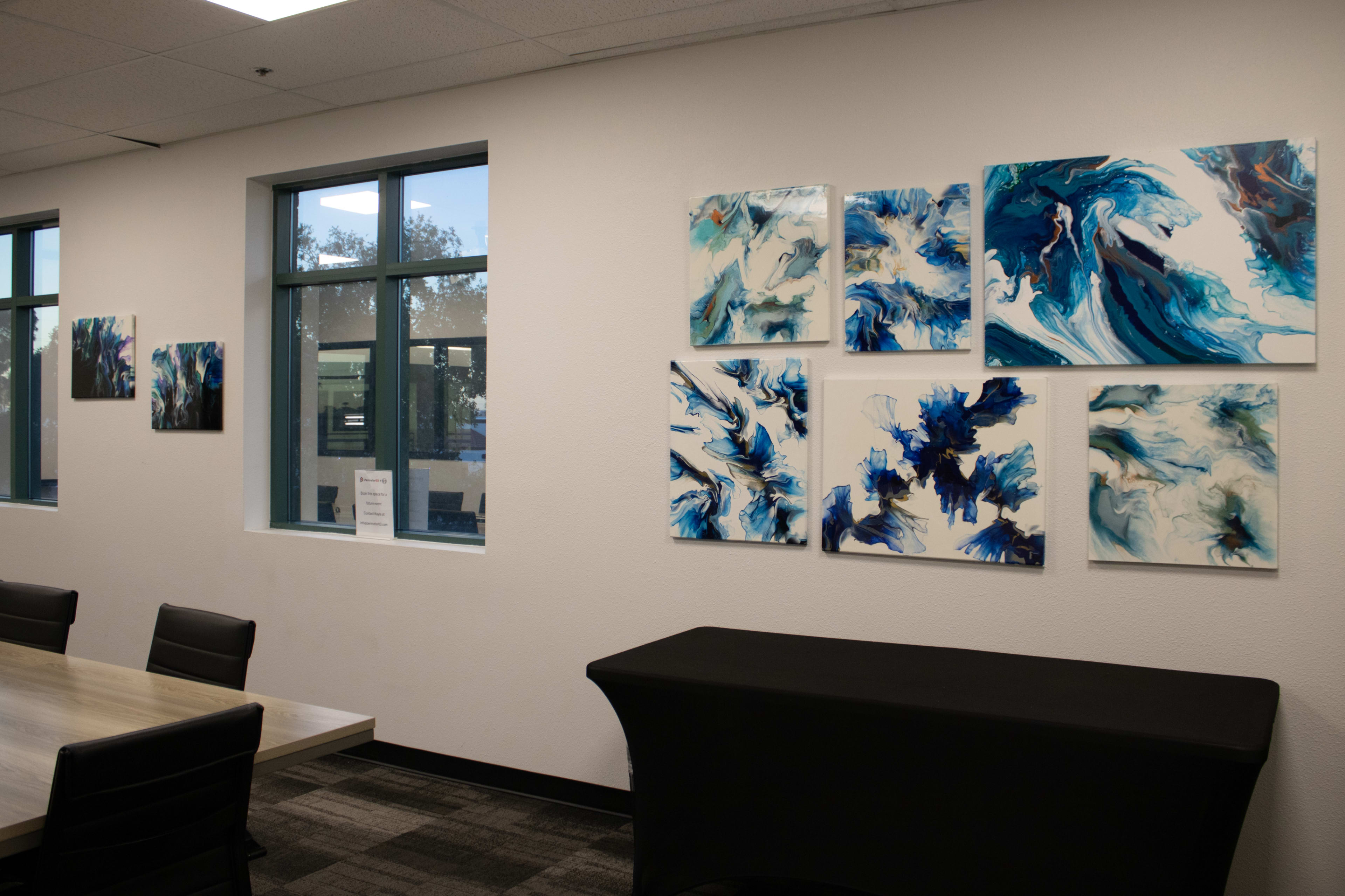 The image shows a modern office space with a long table and black chairs, featuring an arrangement of blue and white abstract art on the walls.