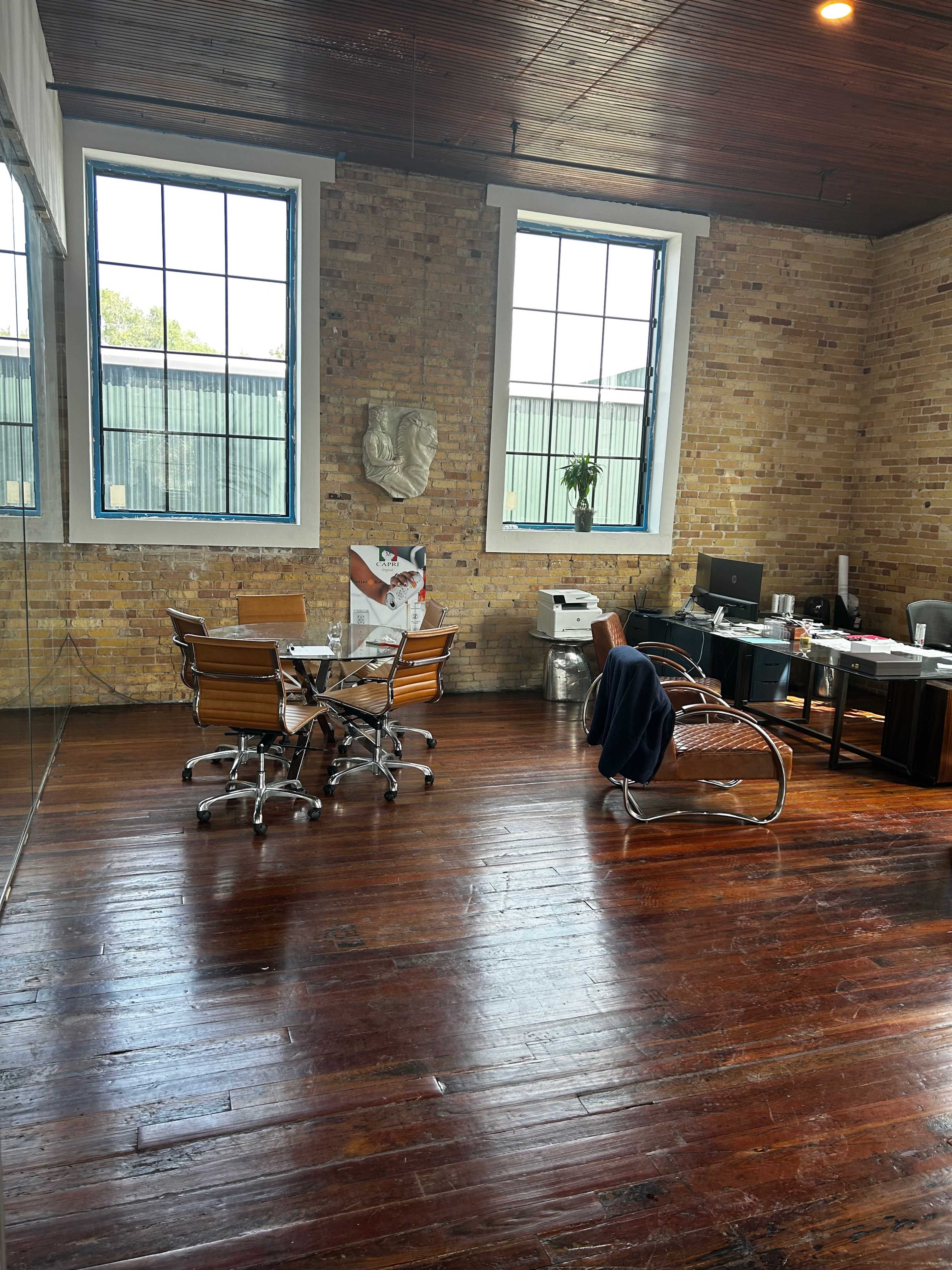 A spacious office with wooden floors, large windows, a meeting table with chairs, and a desk area in the corner.