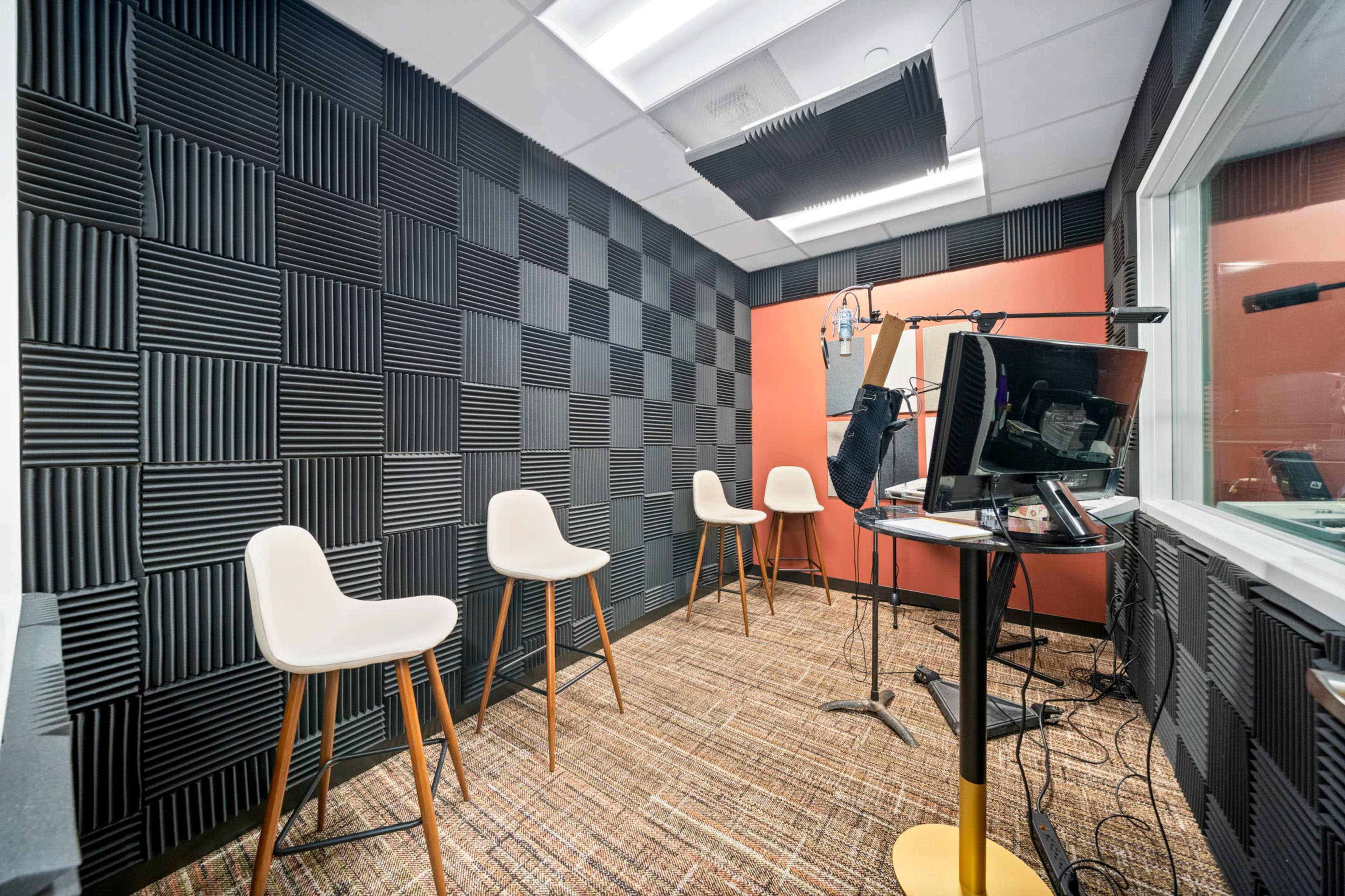 The image shows a soundproof recording studio featuring four white bar stools, a microphone setup, and a monitor, with a wall covered in black acoustic tiles.