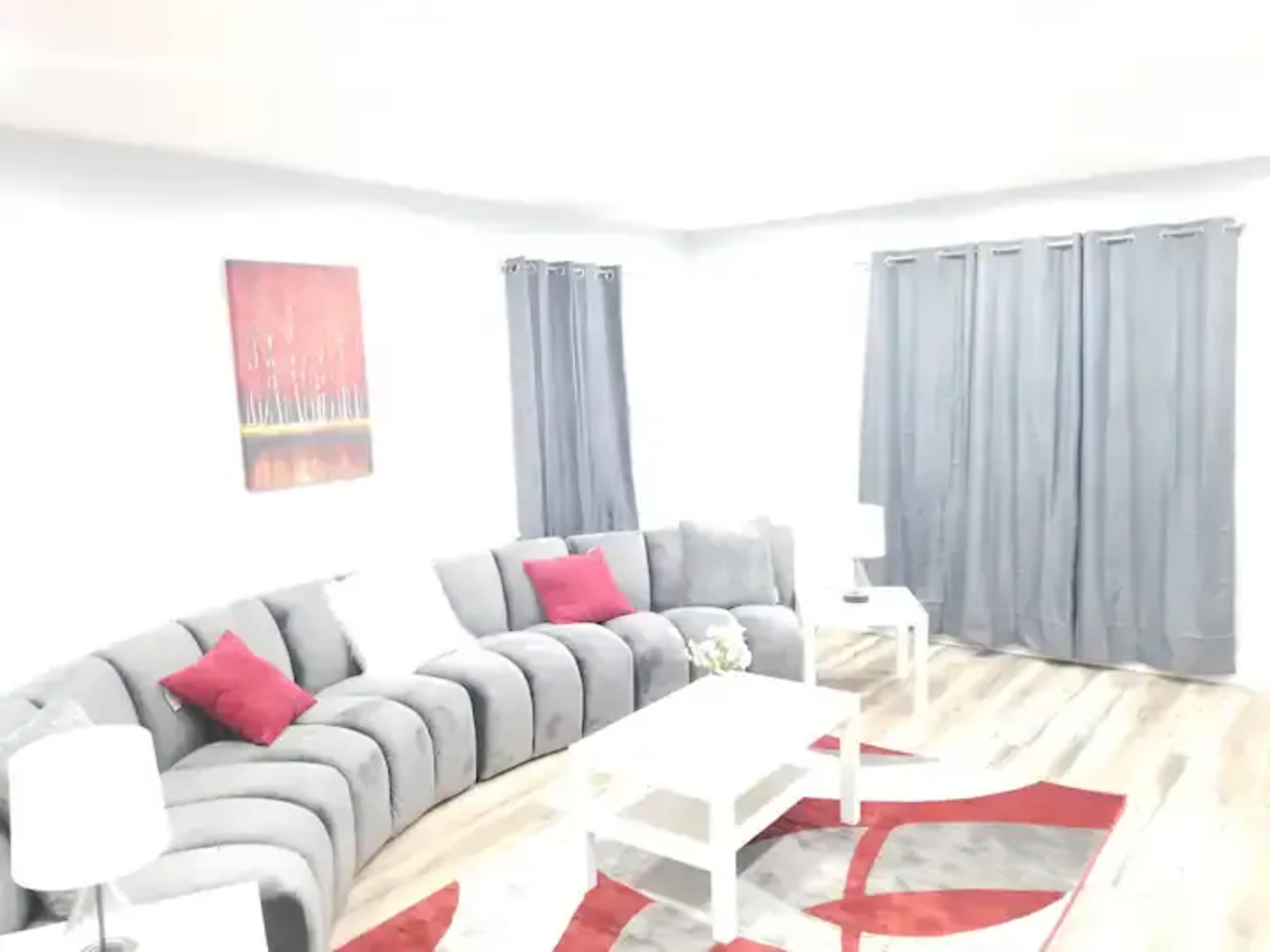 A modern living room features a curved gray sectional couch with red accent pillows, a white coffee table, and gray curtains covering the windows.
