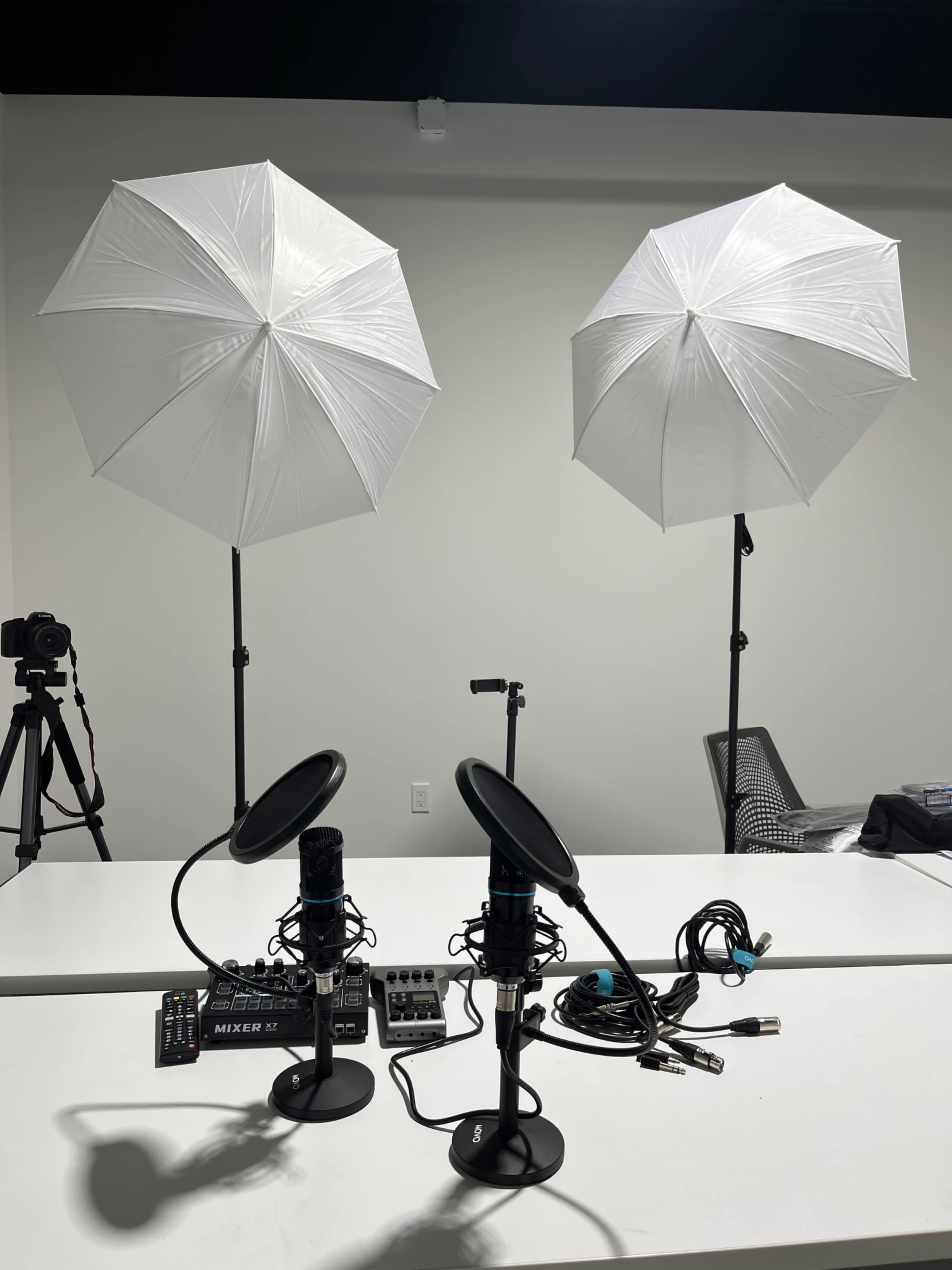 The image shows two studio microphones on a table, flanked by two large white umbrella lights, with audio equipment and cables scattered around.