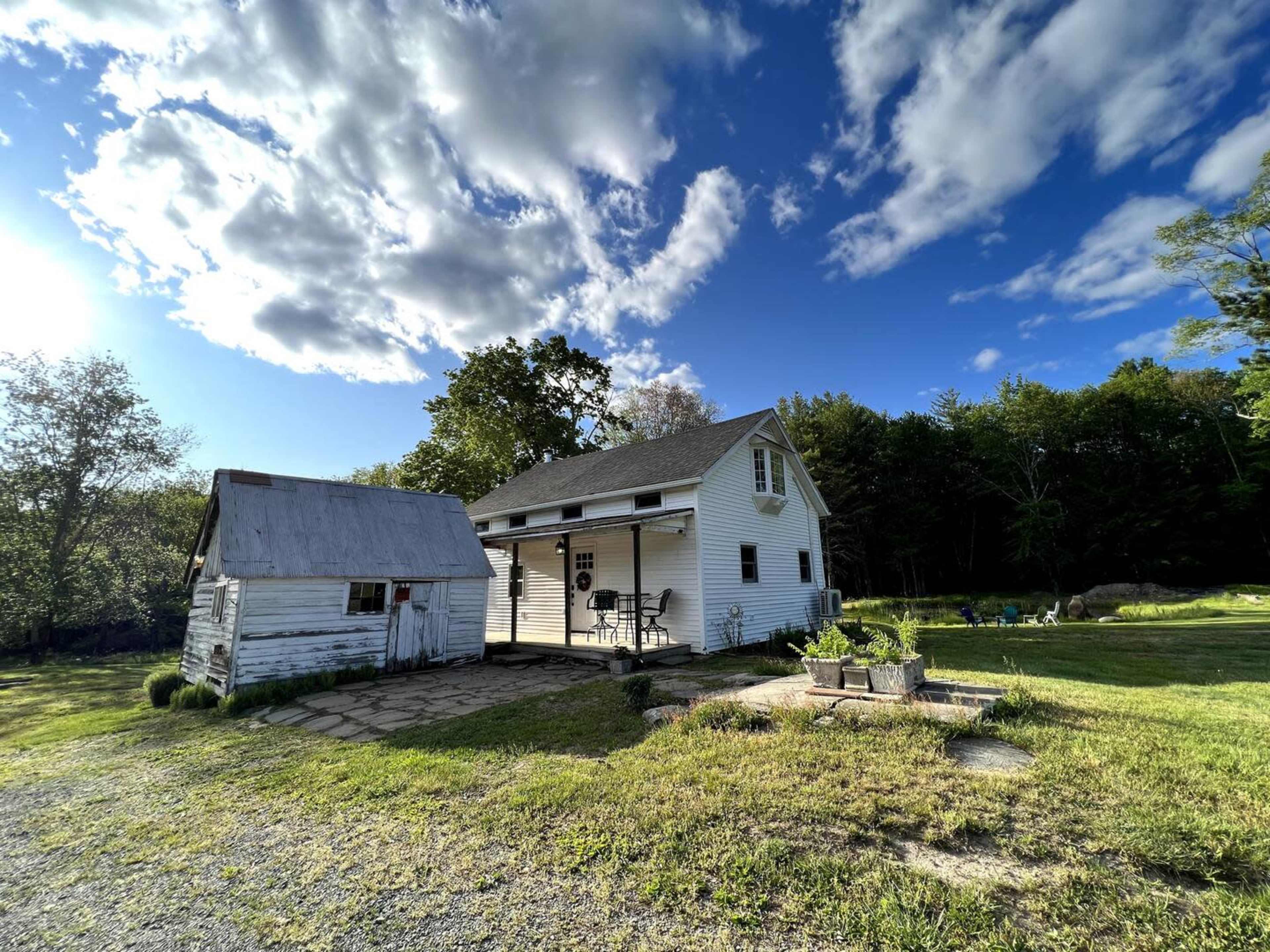 A white farmhouse with a sloped roof stands next to a small shed on a grassy area under a partly cloudy sky.
