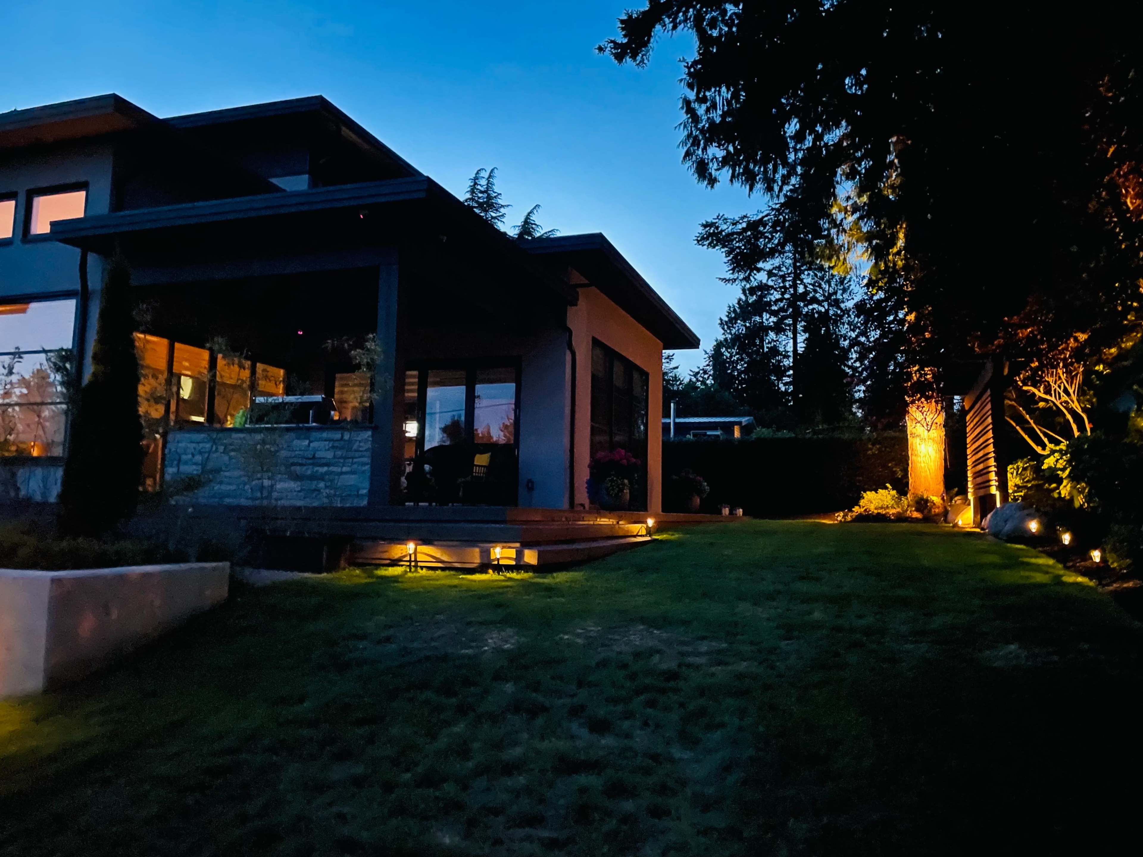 A modern house with outdoor lighting illuminating the garden and patio area during dusk.