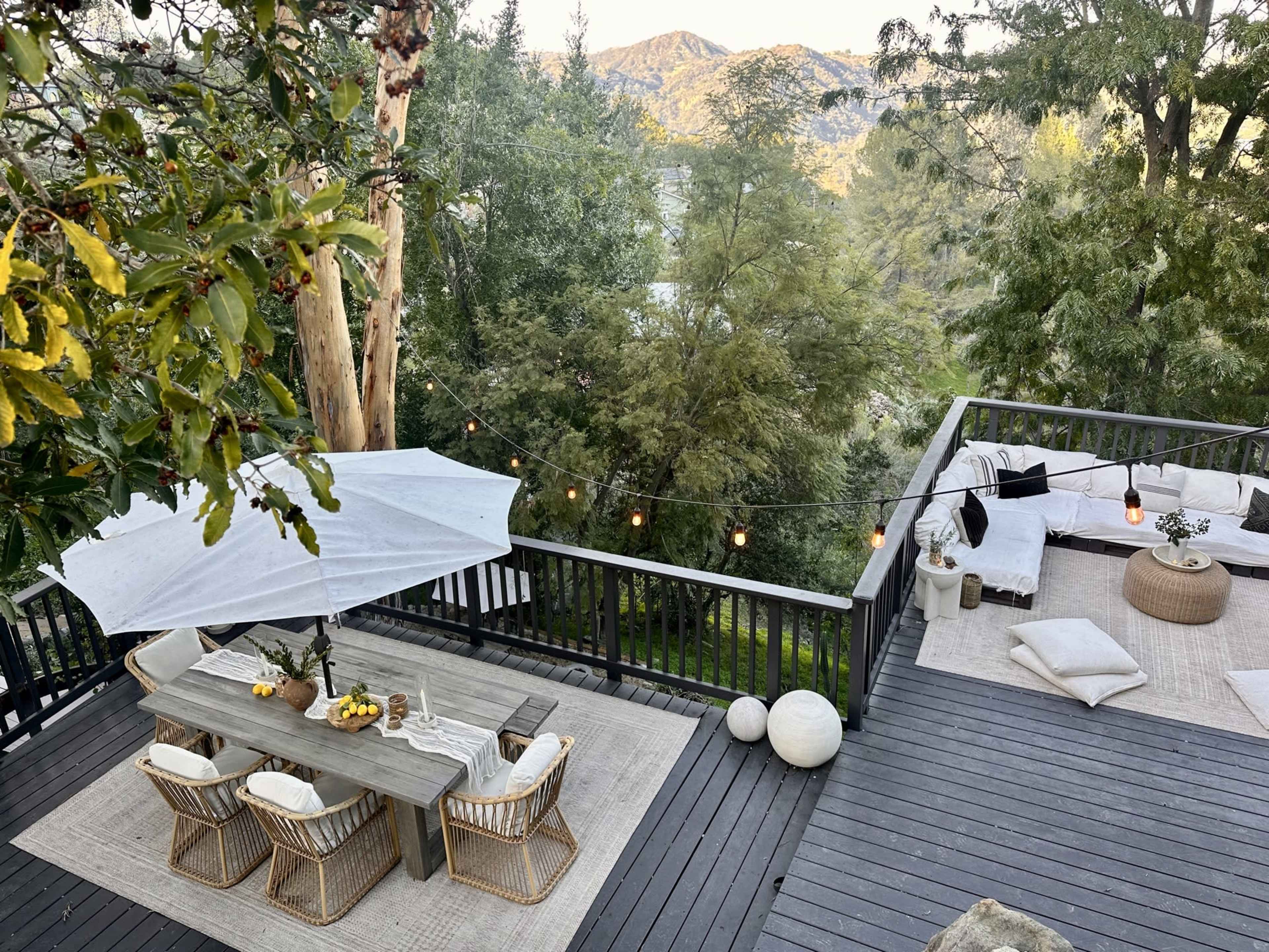 A spacious outdoor deck with a dining table, lounge area, and string lights, surrounded by trees and a view of distant mountains.