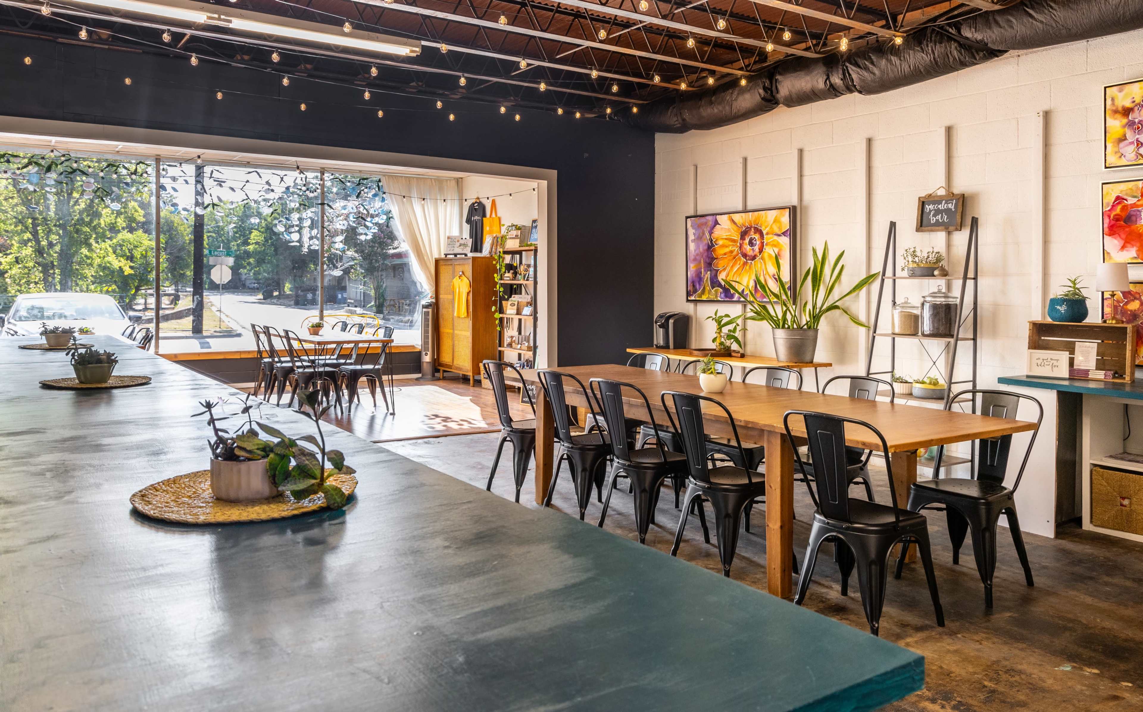 The image shows a bright, spacious interior of a café with wooden tables, industrial-style chairs, and large windows allowing natural light to enter.