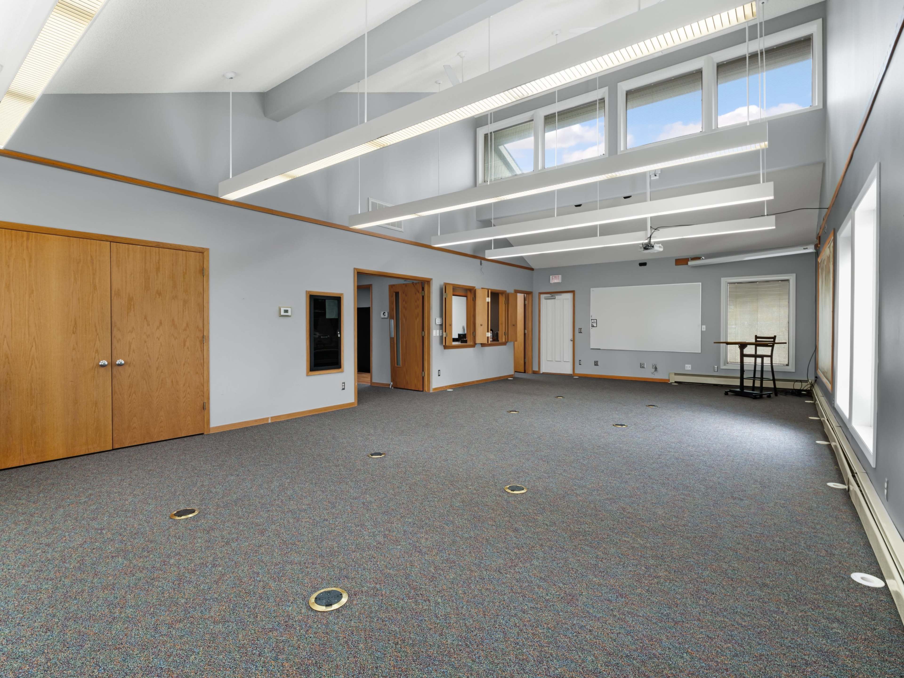 An empty room with gray walls and carpet, featuring large windows, ceiling lights, and several doors.