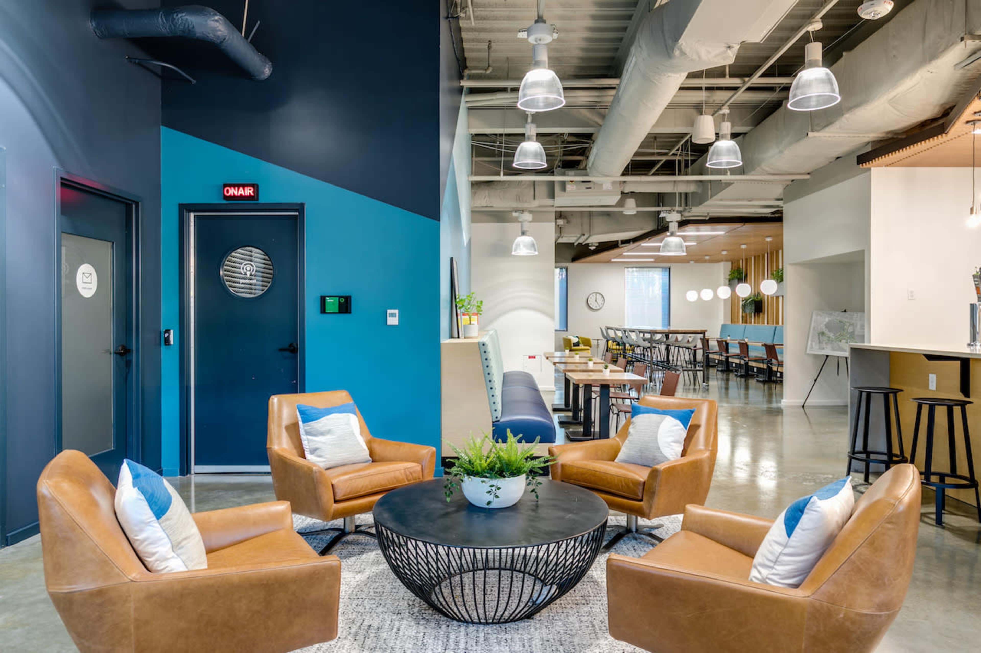A modern lounge area with four brown leather chairs arranged around a circular black table, situated beneath industrial-style lighting and adjacent to a dining space.