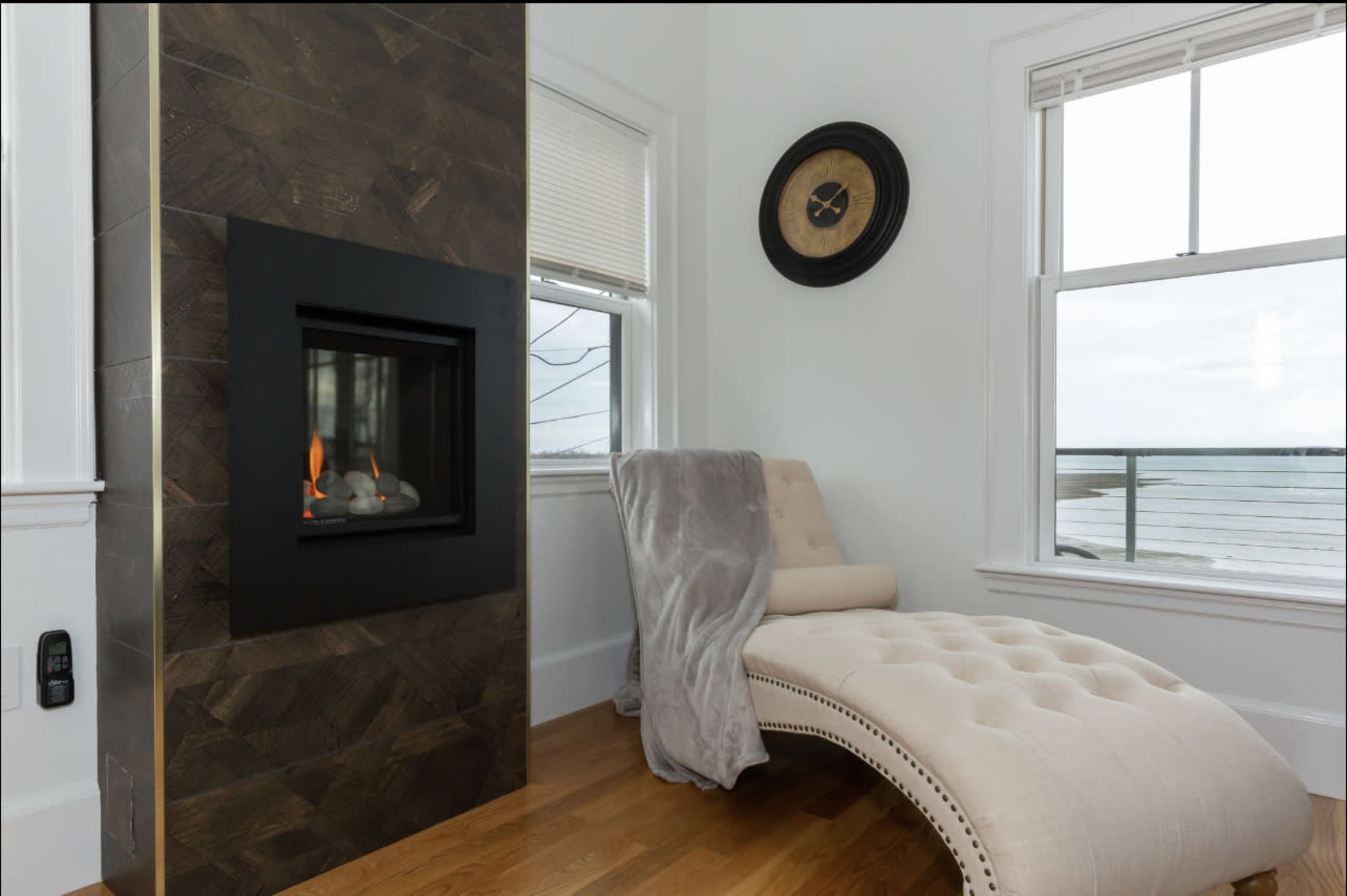 A modern living space featuring a curved chaise lounge next to a black-framed gas fireplace, with large windows offering a view of the outside.
