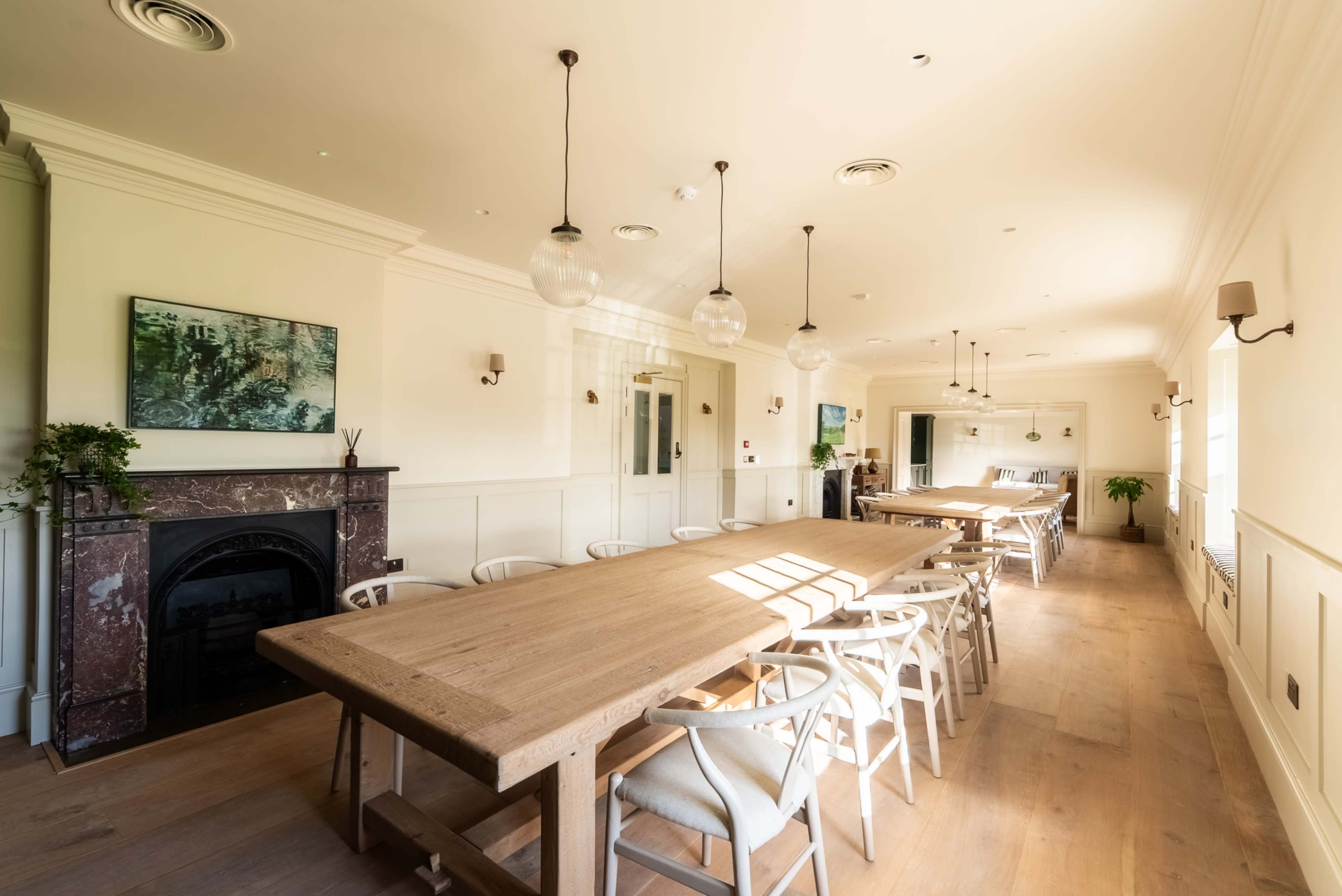 The image shows a spacious dining room with two large wooden tables, surrounded by white chairs, and features large windows allowing natural light to enter.