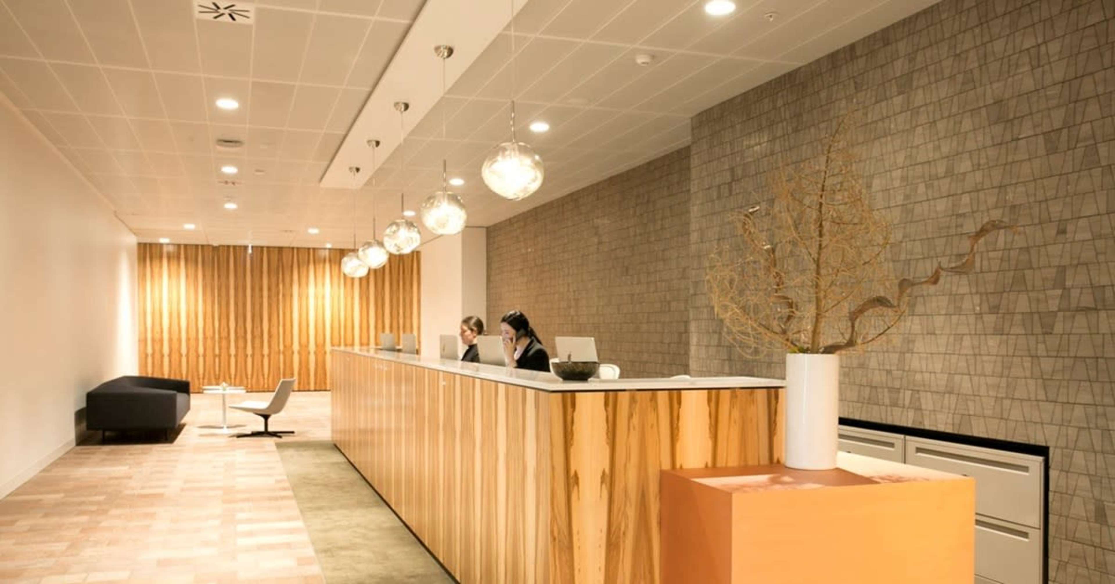 A modern reception area with a wooden desk, two staff members seated behind it, and pendant lighting overhead.