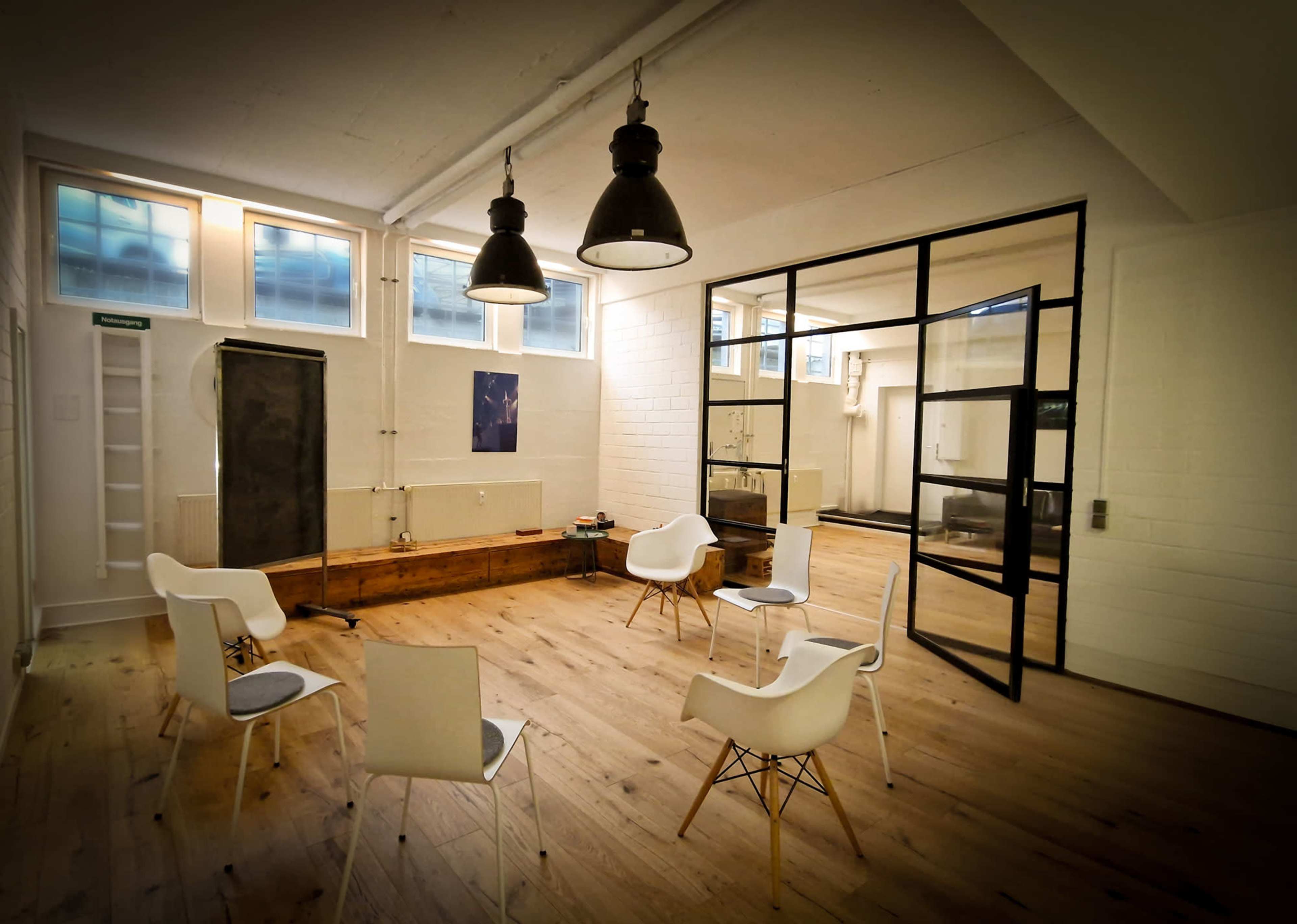 The image shows a modern, minimalist meeting room with white chairs arranged in a circle on a wooden floor, illuminated by pendant lights.