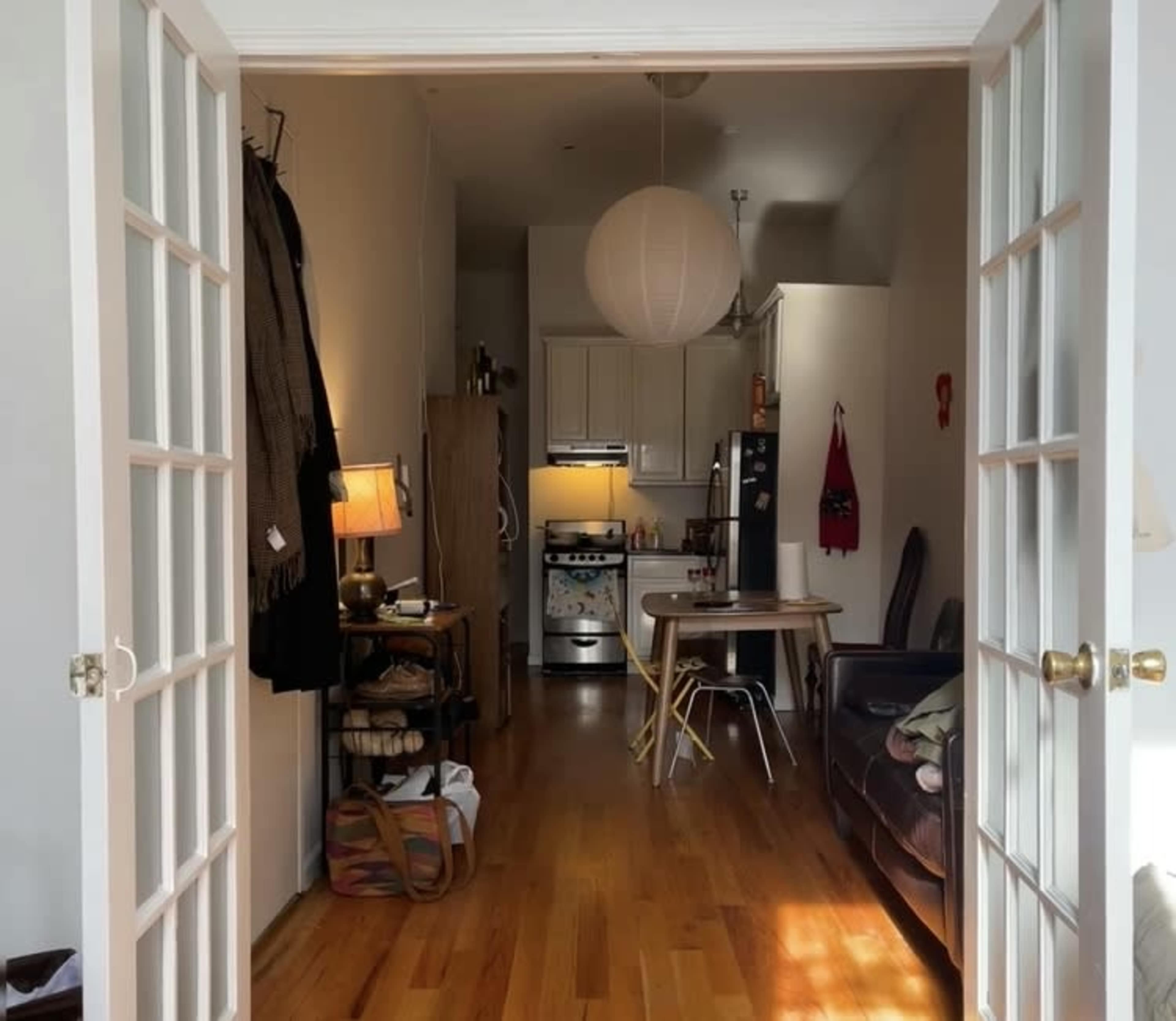 A view through a set of French doors reveals a cozy living space with a table and chairs, an open kitchen area, and wooden flooring.